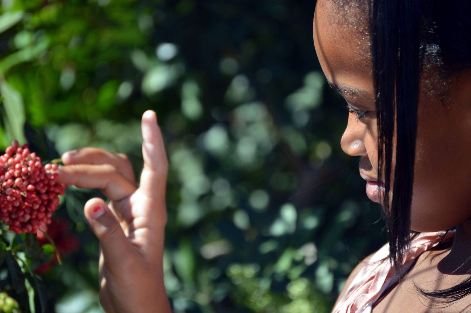 Young girl closely examining a vibrant red flower cluster in a lush green garden setting.