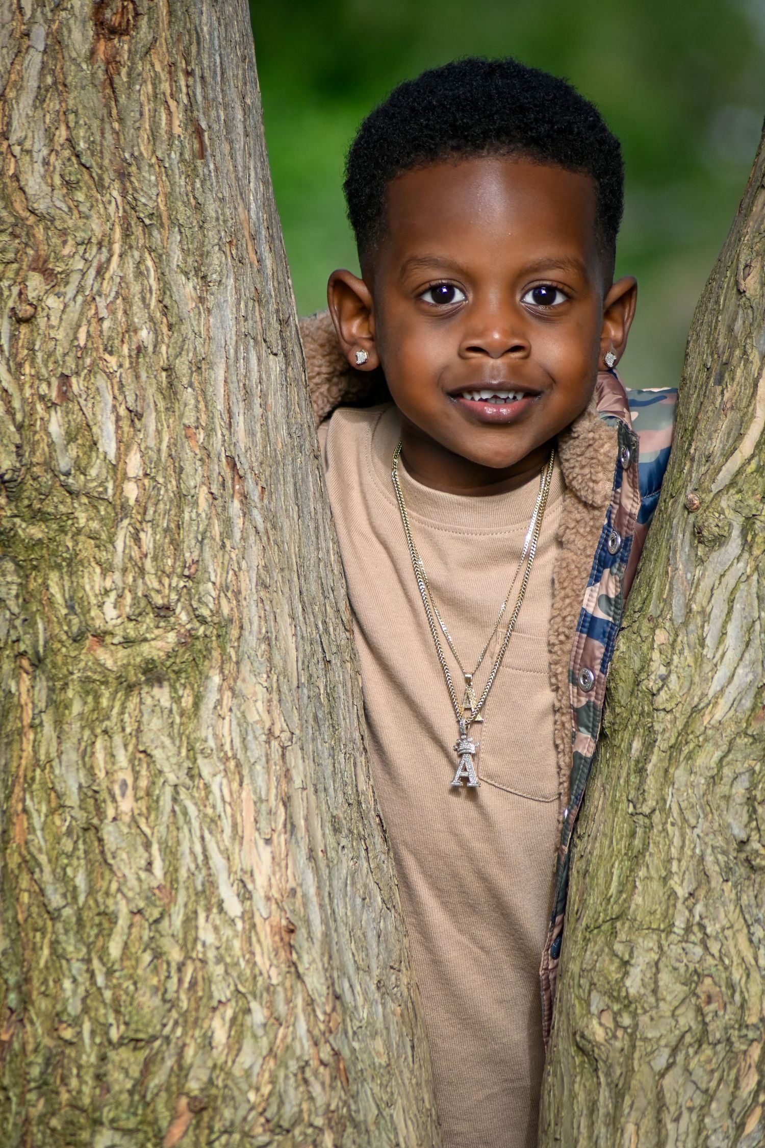 A young Black boy smiles between two tree trunks, wearing a tan shirt and necklace outdoors.