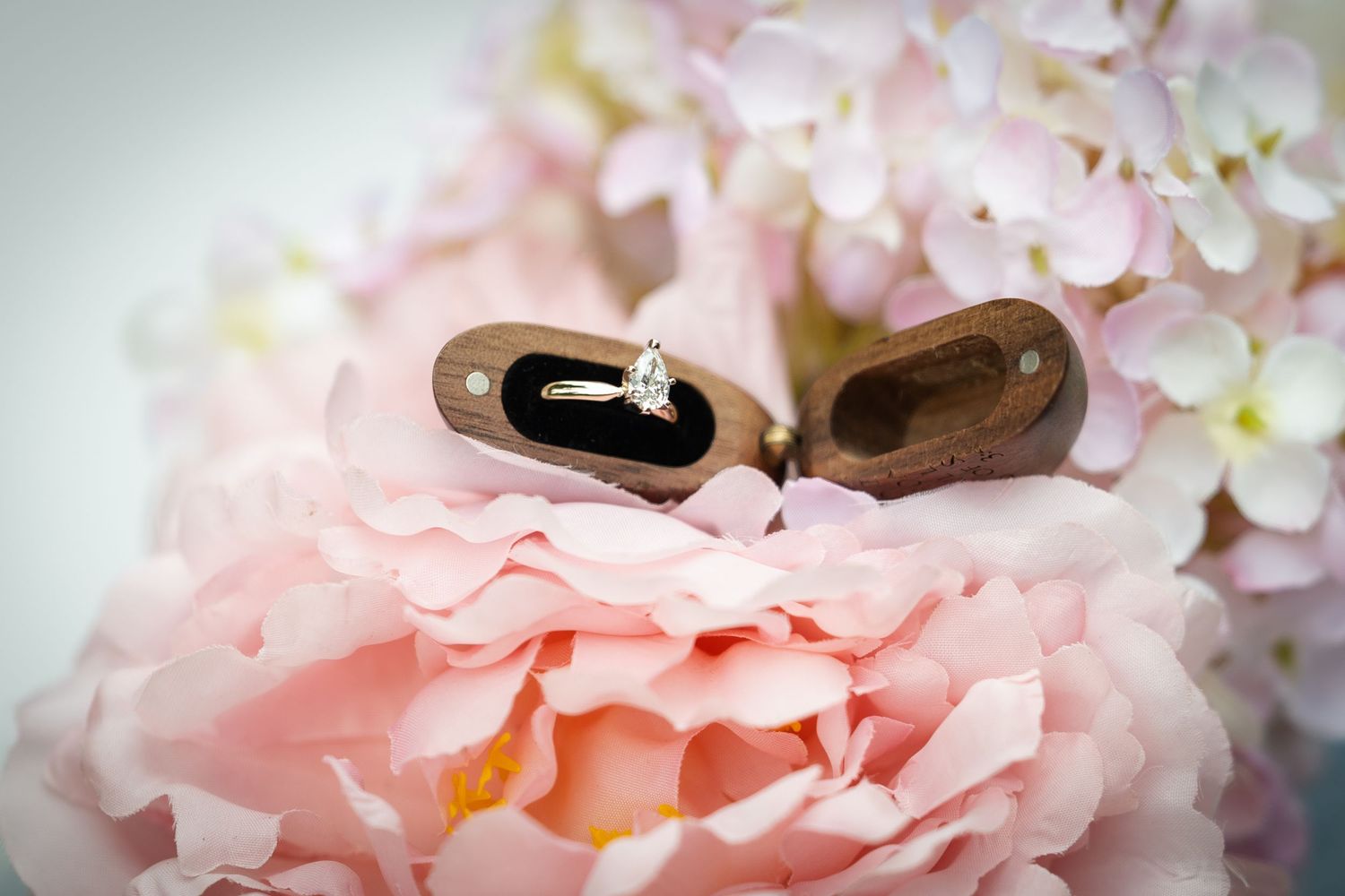 Diamond engagement ring in wooden heart-shaped box resting on pink peonies and hydrangeas.