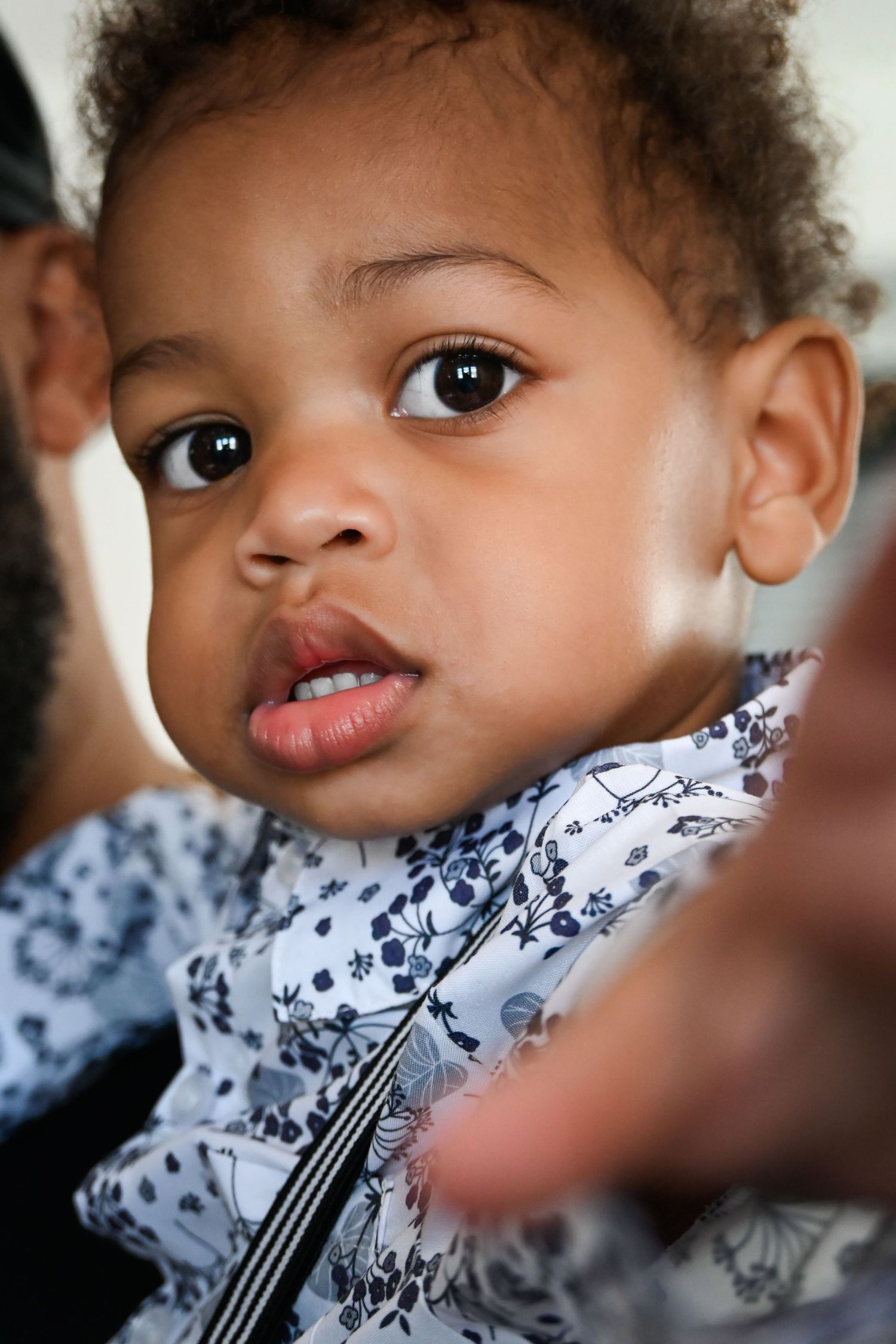 Adorable Black baby with curly hair and big brown eyes wearing a floral outfit, held by an adult.