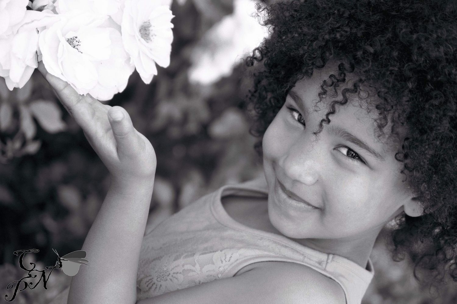 Black and white portrait of a young girl with curly hair smiling while holding a white flower.