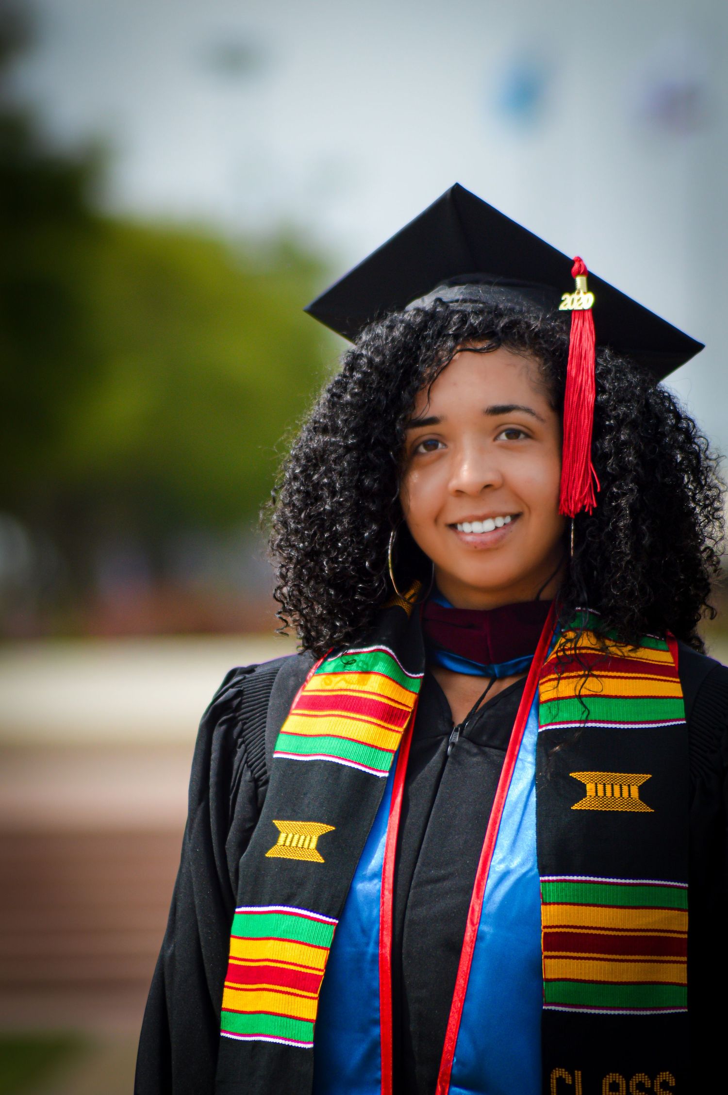 Graduate wearing black cap and gown with colorful kente stole smiling during graduation photo session outdoors.