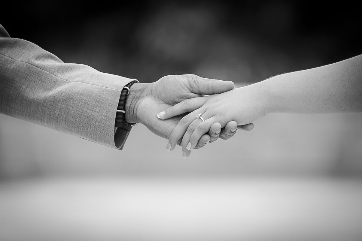 Black and white photo of a couple holding hands, showing a wedding ring and bracelet in a romantic close-up.