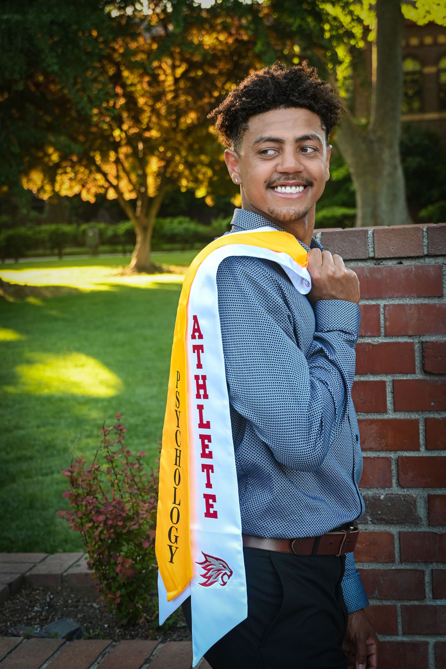 A graduate in a blue sweater poses against a brick wall wearing a graduation sash with school colors and text.