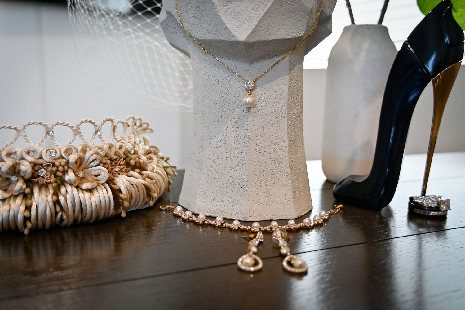 Gold pearl necklace draped on white geometric vase beside bridal accessories and black stiletto heel on dark wood surface.