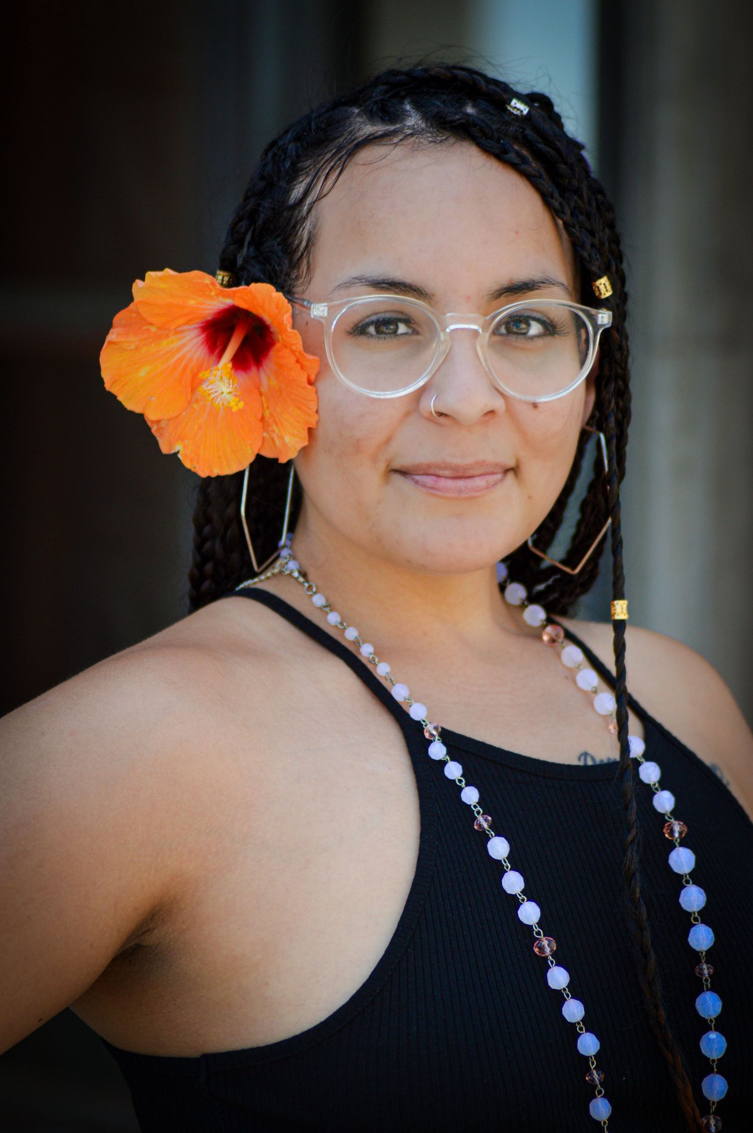 Portrait with clear glasses wearing a black tank top, beaded necklace and vibrant orange hibiscus flower in dark hair.