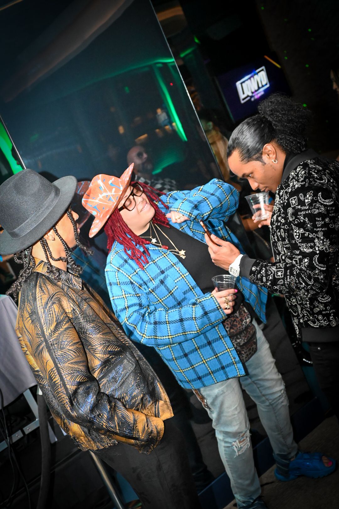 People socializing at a nightclub with green lighting and casual attire including plaid shirts and hats.
