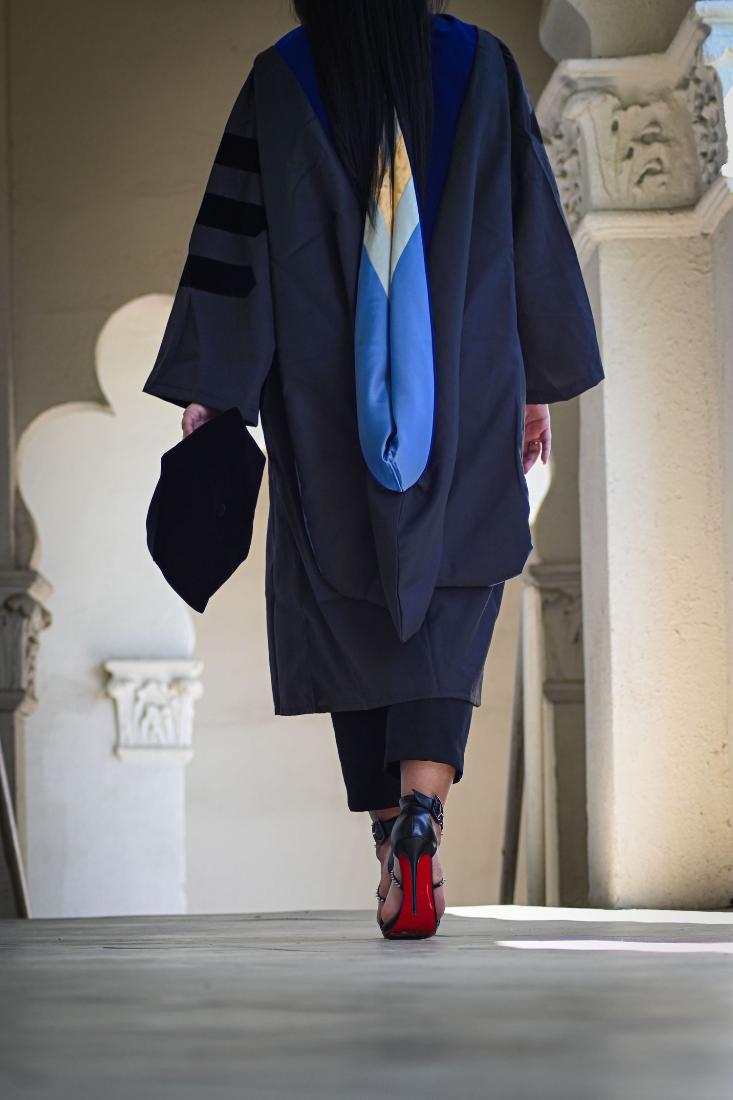 Low angle view of graduate walking in academic regalia showing blue hood and red-soled shoes.