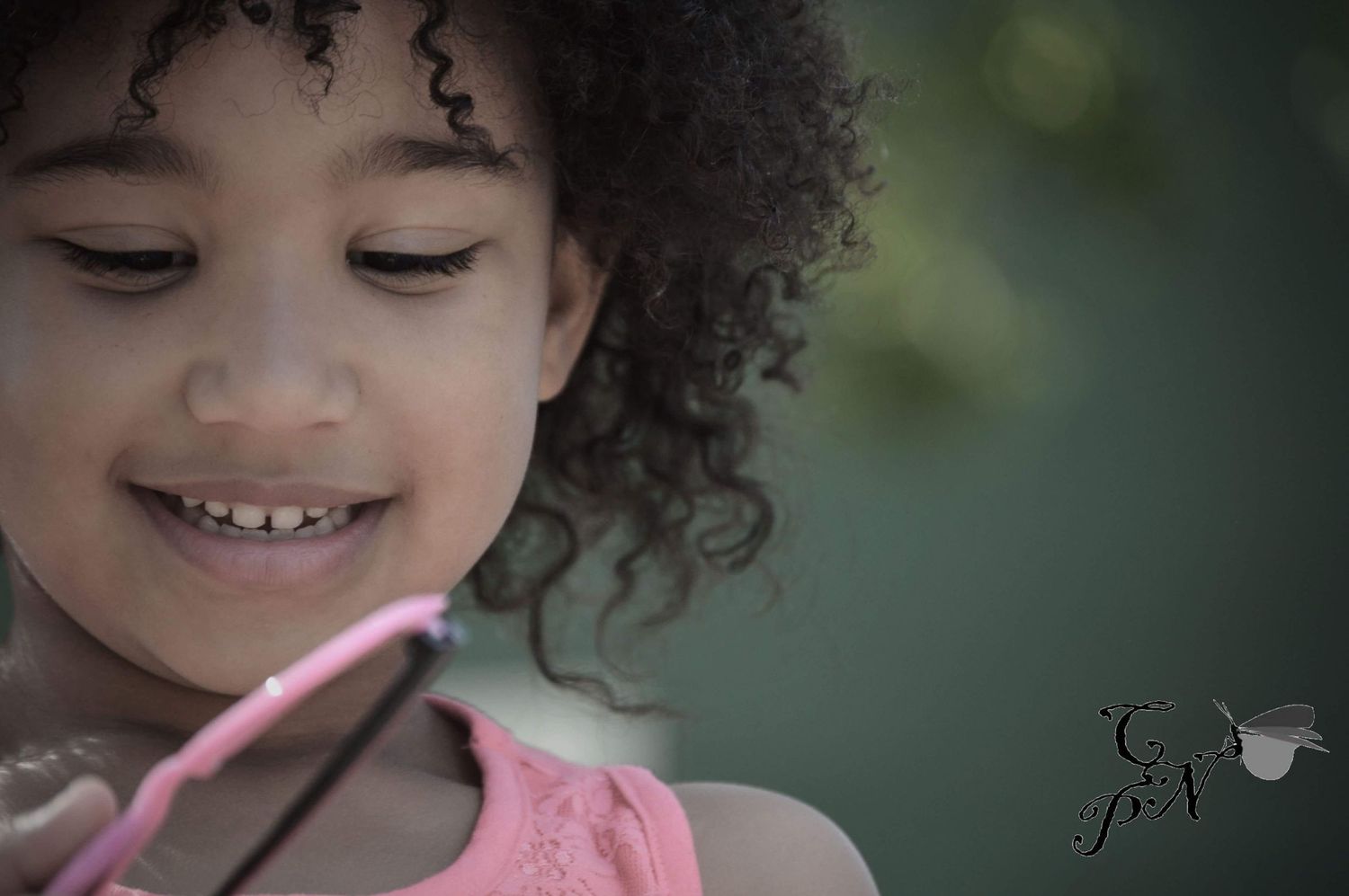 Young girl with curly hair smiling while holding a pink object outdoors in natural light.