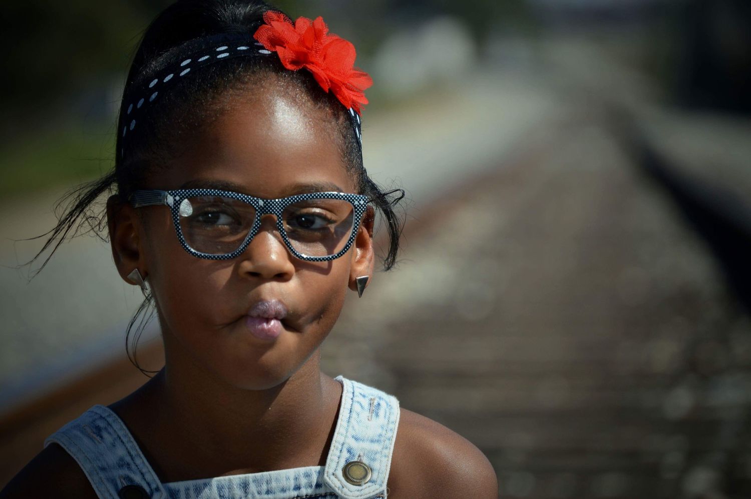Young girl with red flower hair accessory and blue glasses standing near railroad tracks outdoors.