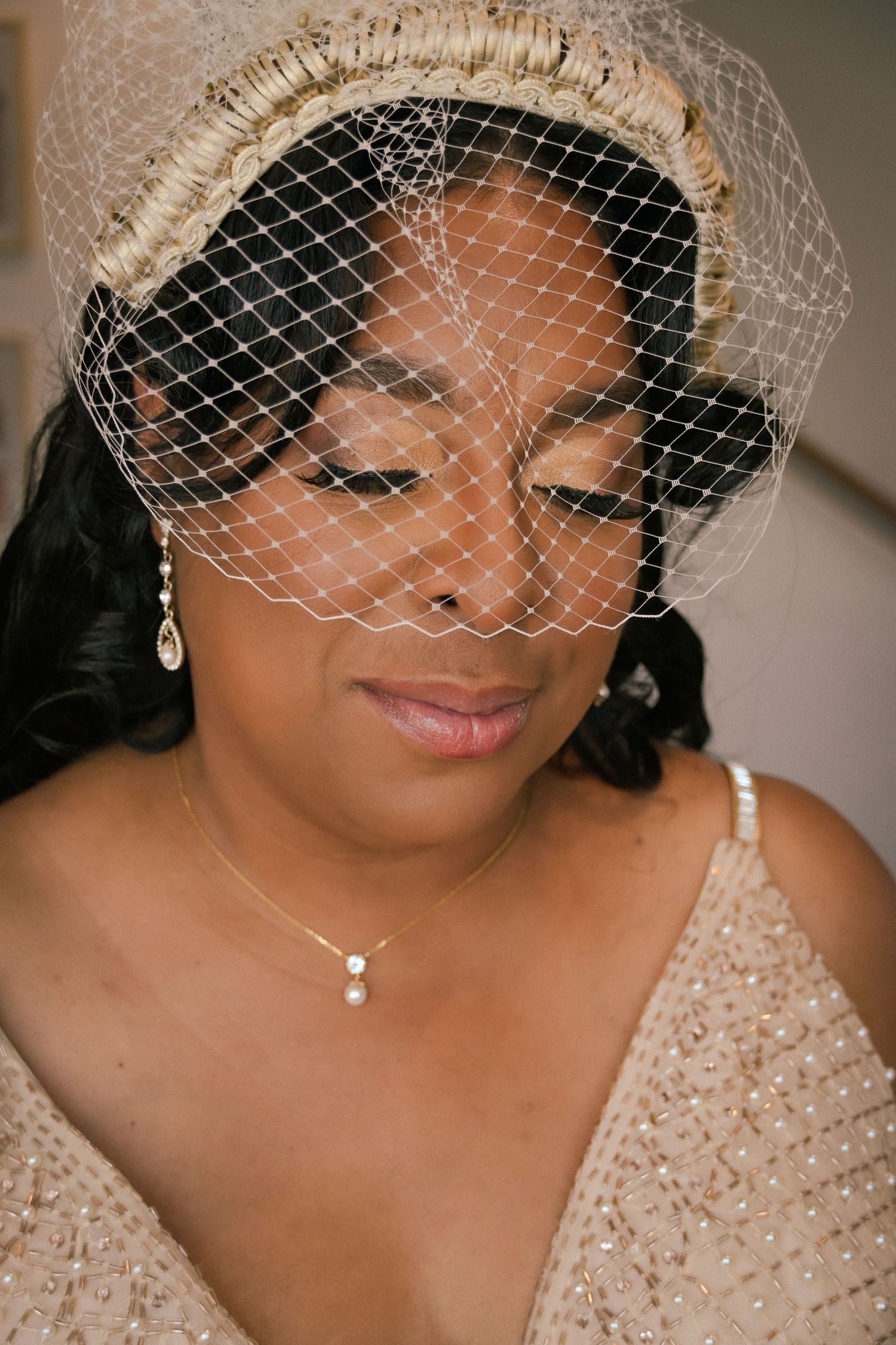 Elegant bride wearing a birdcage veil and pearl jewelry, looking down gracefully in a beaded gown.