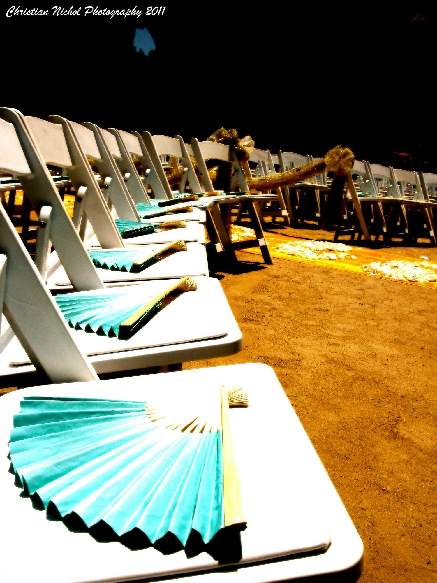 Rows of white folding chairs with teal paper fans on seats arranged on sandy ground for an outdoor wedding ceremony at night.
