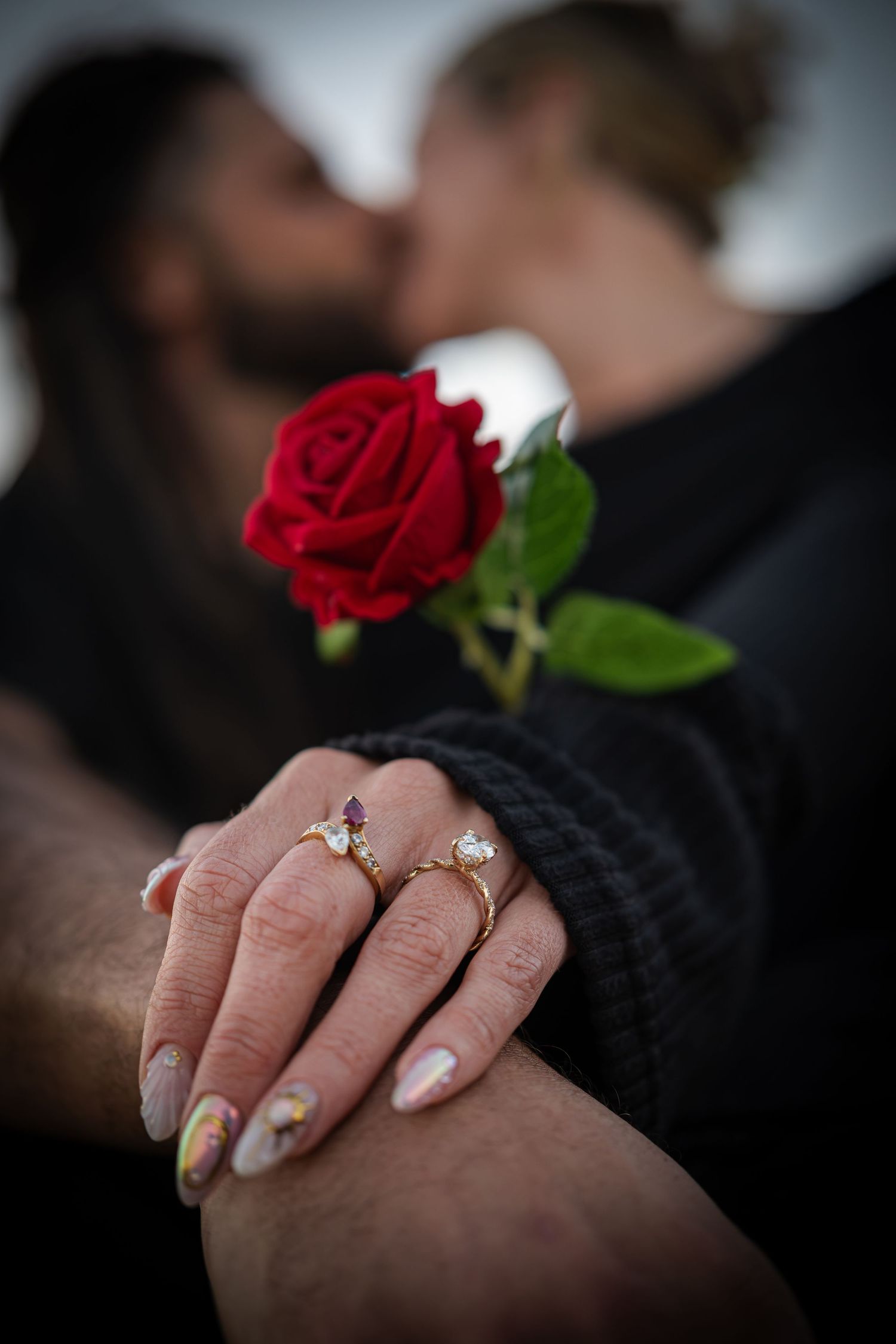 Couple kissing while holding a red rose, with gold engagement rings visible on the woman's hand in the foreground.
