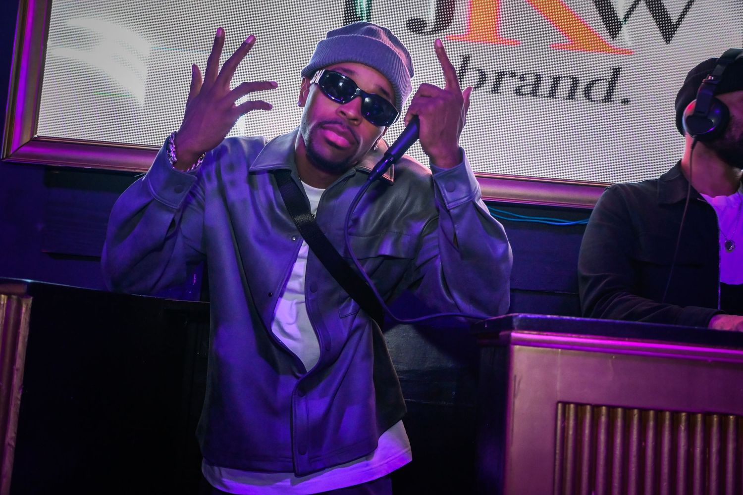 DJ in sunglasses and purple lighting gestures with hands while performing at a nightclub event.