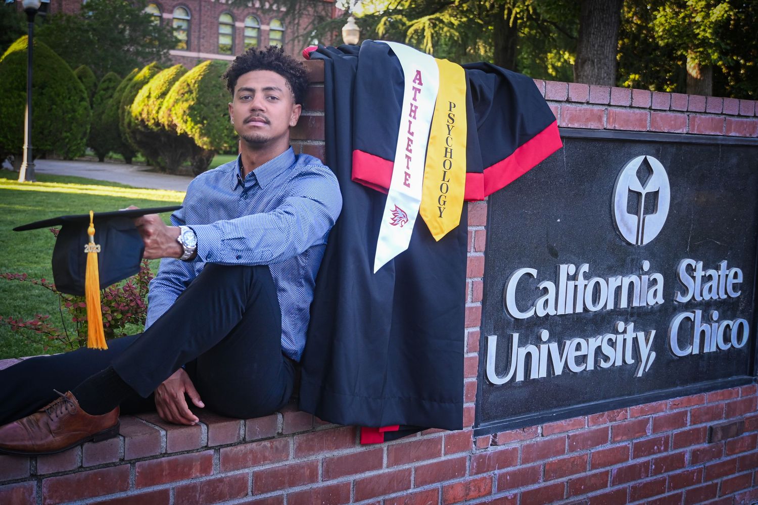 College graduation ceremony with students in caps and gowns celebrating near brick entrance sign and palm trees.