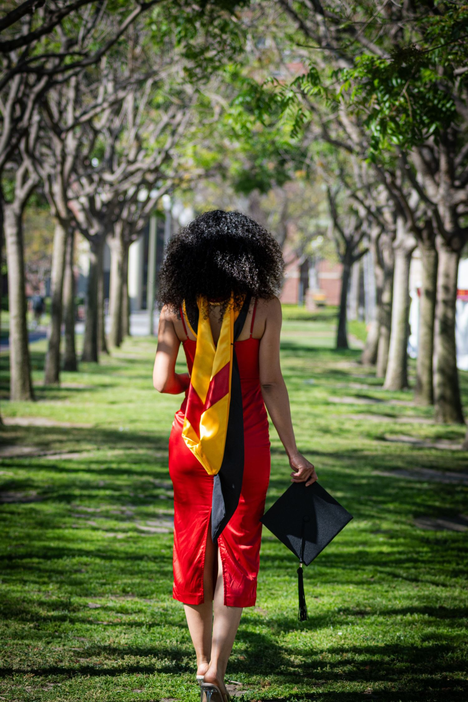Graduate in red regalia walking through a tree-lined path holding a graduation cap.
