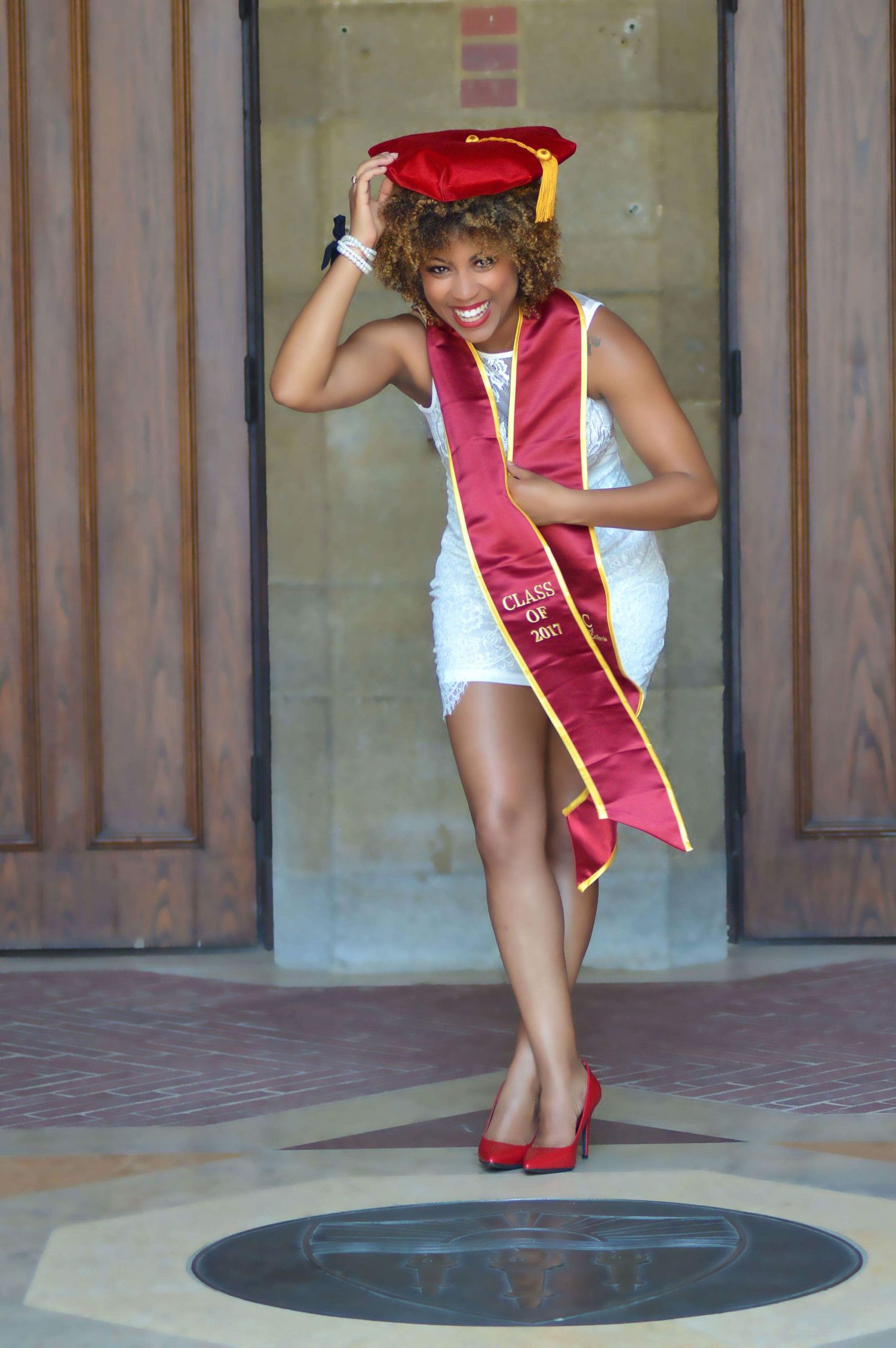 Graduate in white dress and red graduation stole poses playfully with graduation cap.
