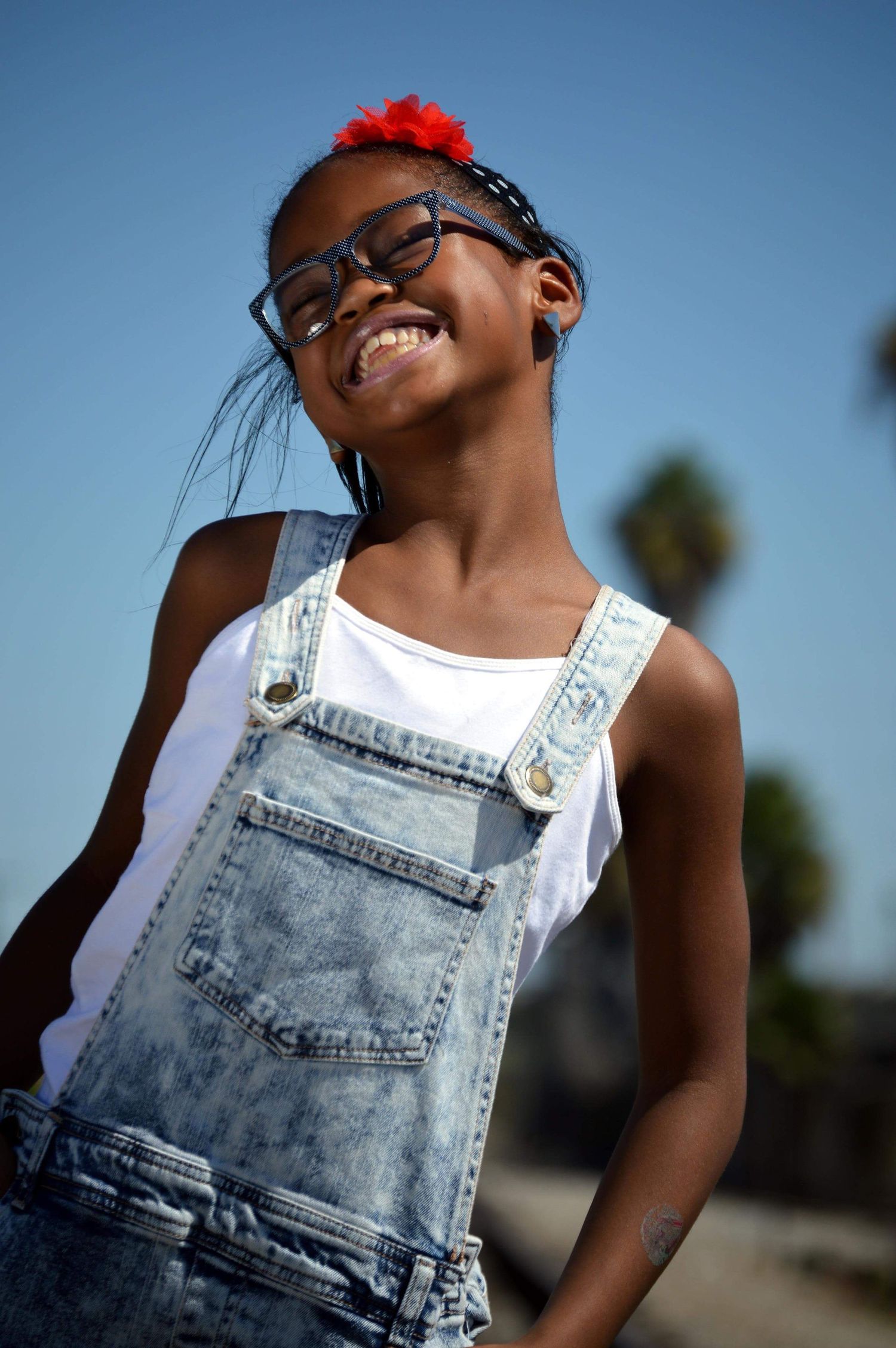 Smiling young Black girl wearing denim overalls, white tank top, and glasses with a red hair accessory outdoors.