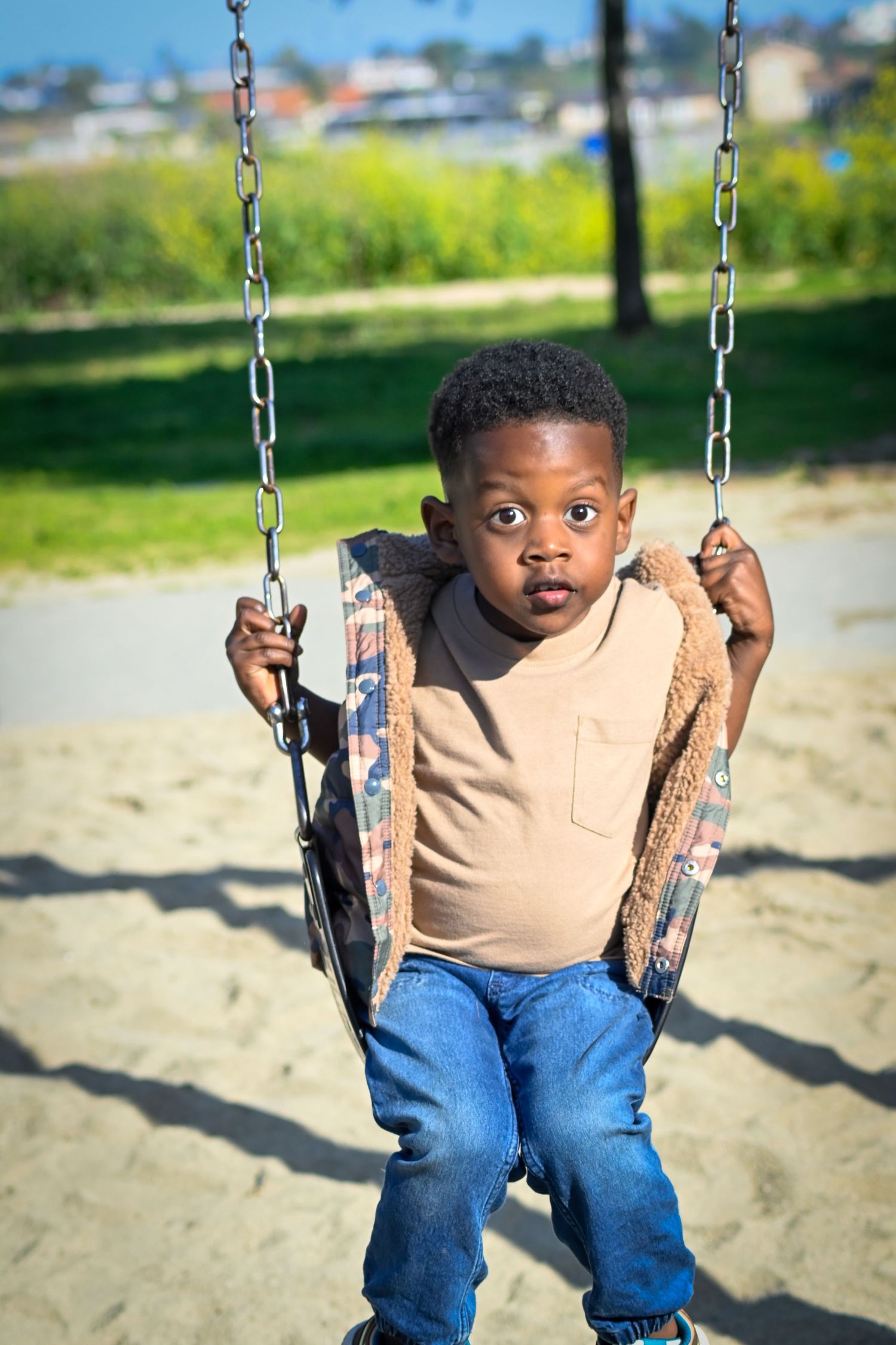 Young Black boy sitting on a playground swing, holding chains, wearing jeans and a tan shirt, looking at camera.