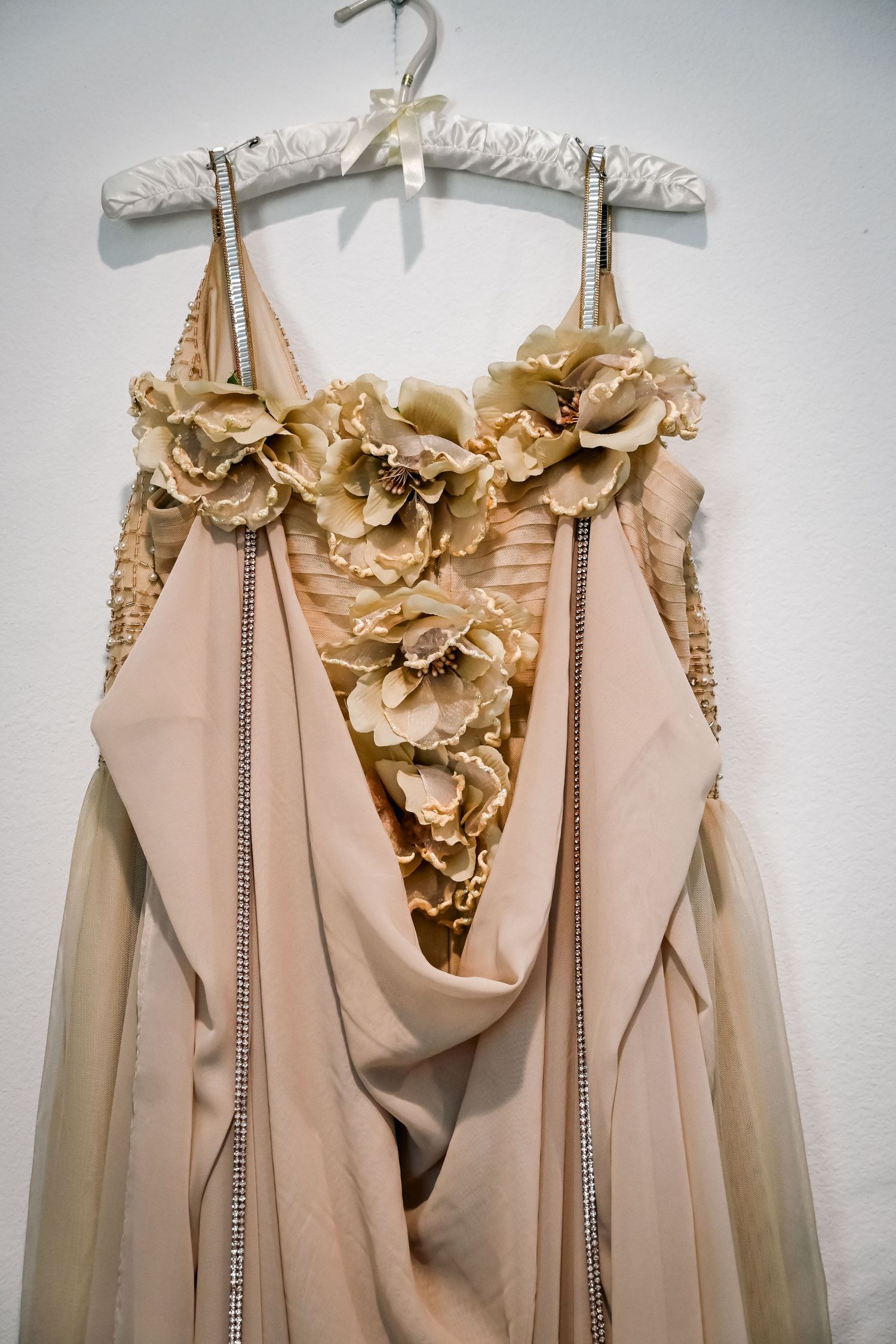 Elegant beige gown with floral embellishments and rhinestone trim displayed on a padded hanger against a white wall.