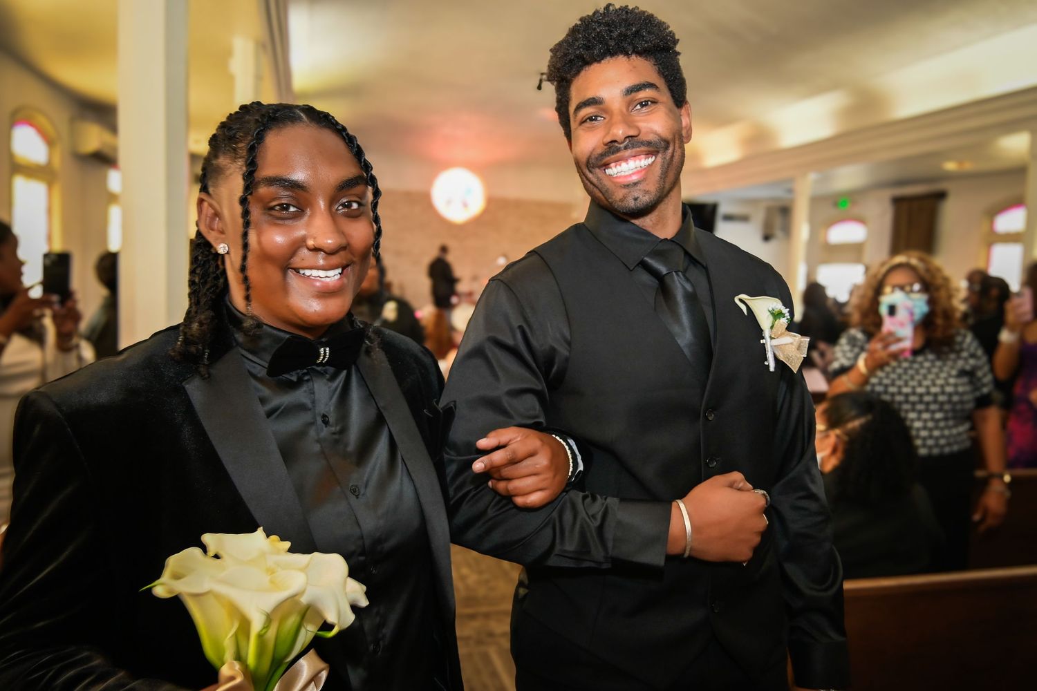 Two people in black formal attire smile at a wedding ceremony, one holding white calla lilies.