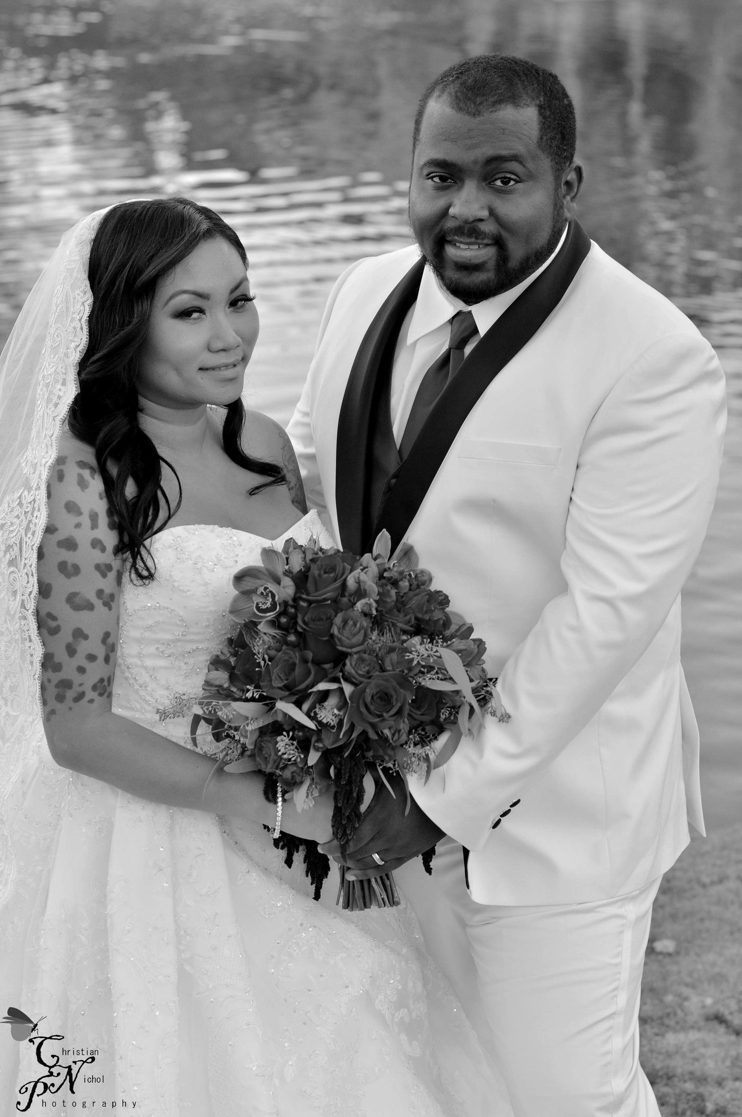 Black and white wedding photo of a bride holding a bouquet and groom in white tuxedo posing near a lake.