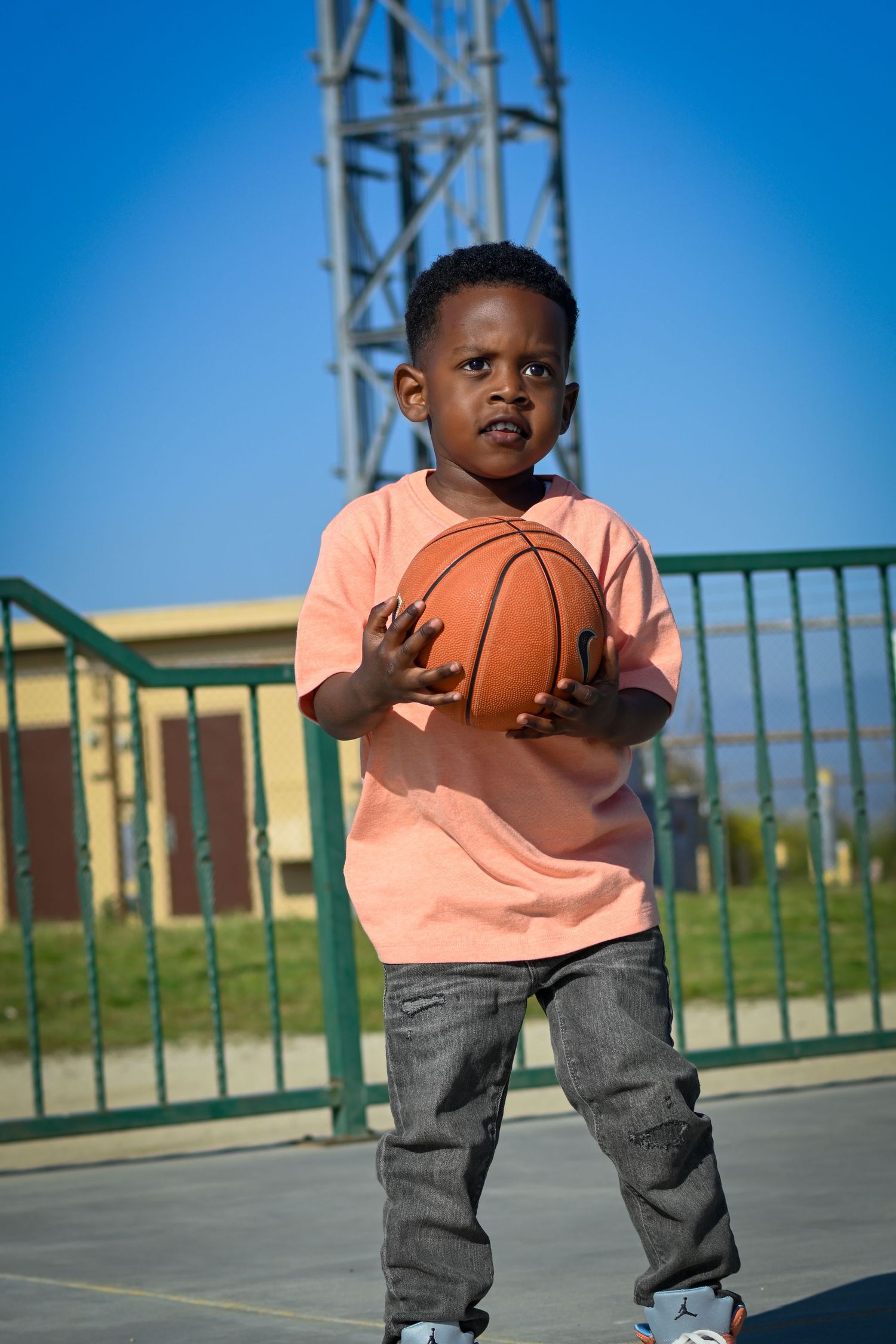 Young Black boy in pink shirt holds basketball on outdoor court with green fence and blue sky background.