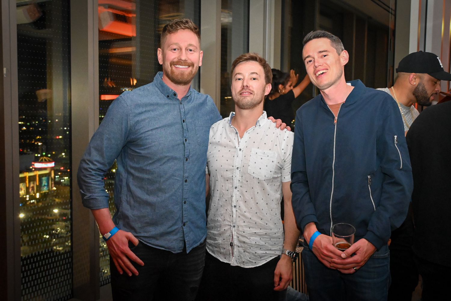Three people pose together at an evening social event with city lights visible through windows in the background.