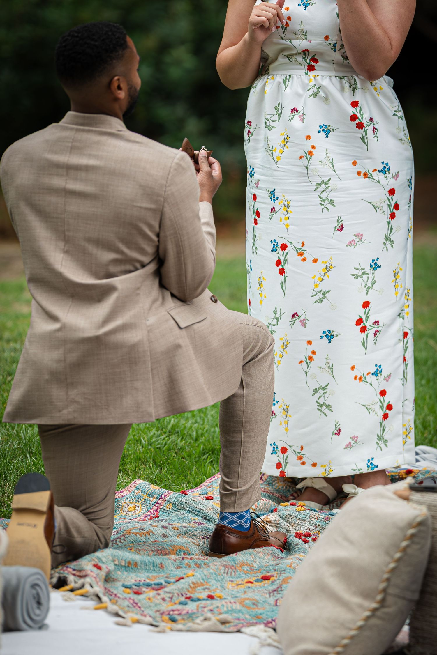 Man in tan suit proposing on one knee to woman in floral dress on a decorated outdoor blanket.