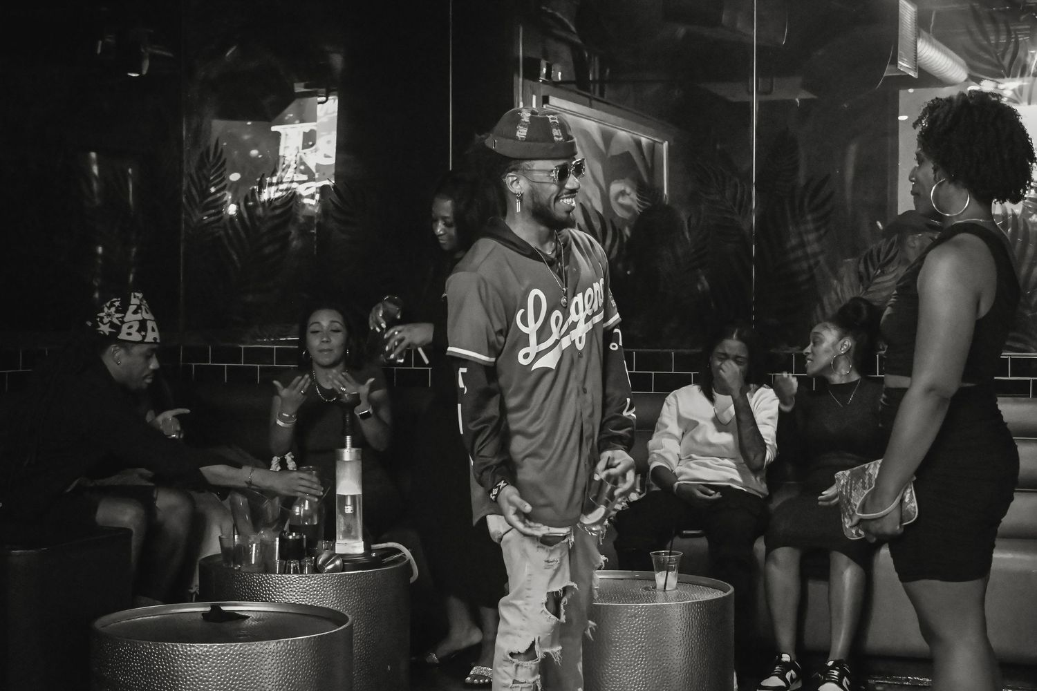 A bartender pours drinks at a dimly lit bar counter while patrons socialize in the background.
