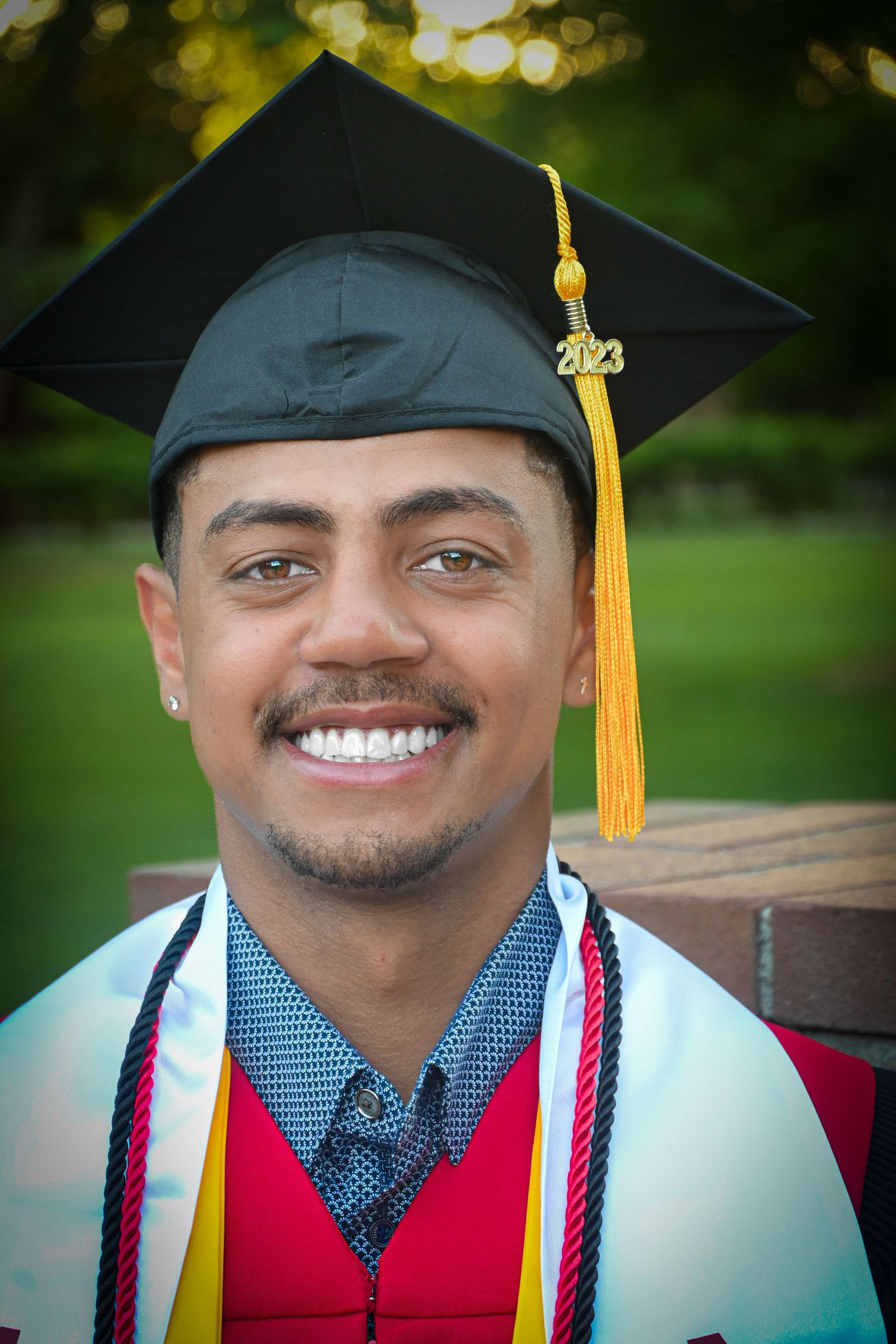 Graduate wearing a polka dot tie, yellow tassel, and graduation stole smiles brightly against a blurred green background.