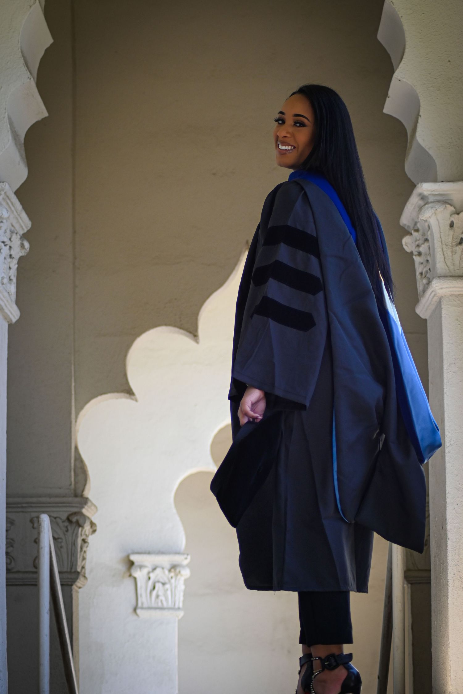 Graduate in dark academic regalia poses in arched hallway with ornate columns.
