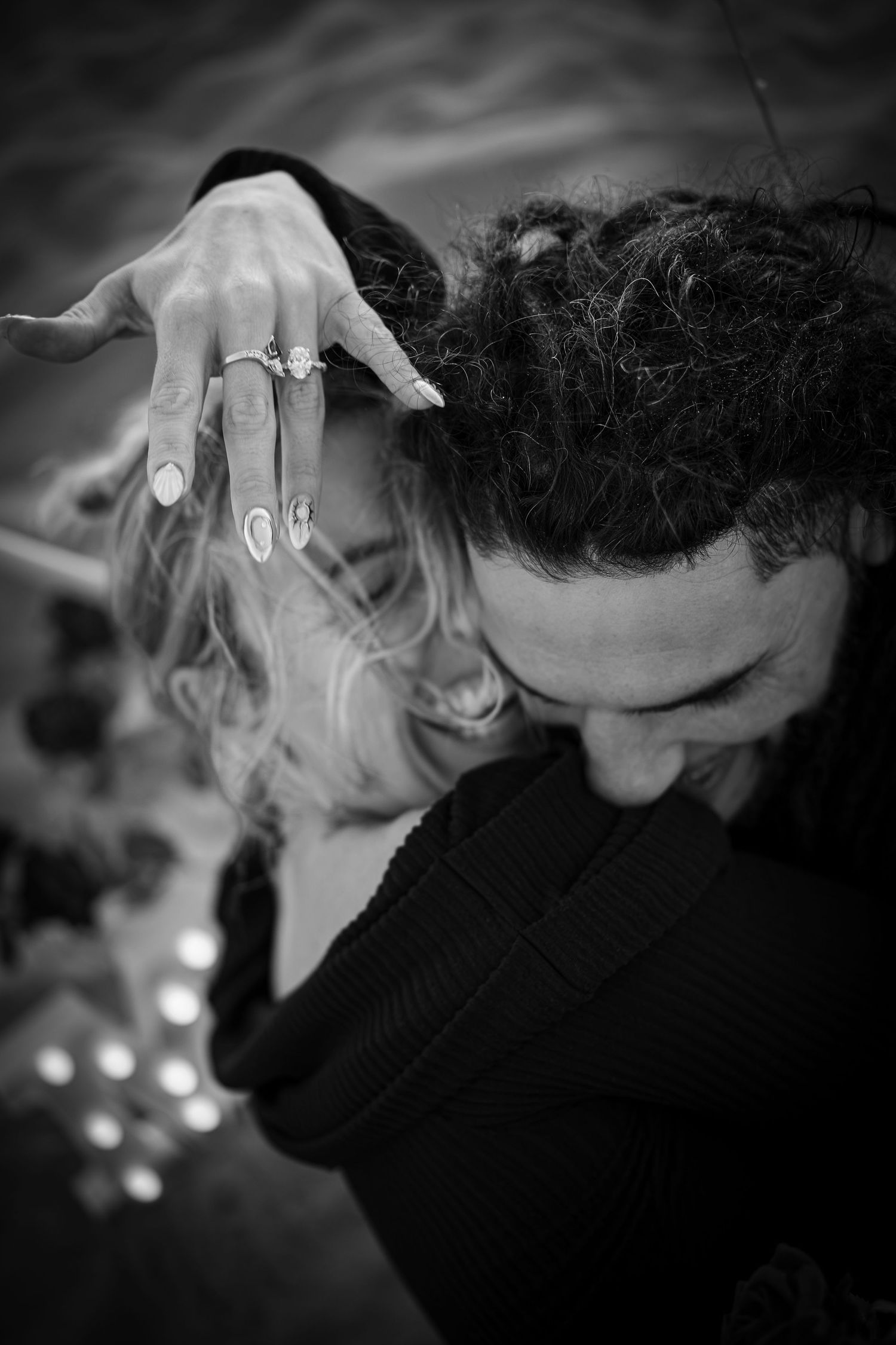 Black and white photo of a couple embracing, woman showing off engagement rings on her hand while smiling joyfully.