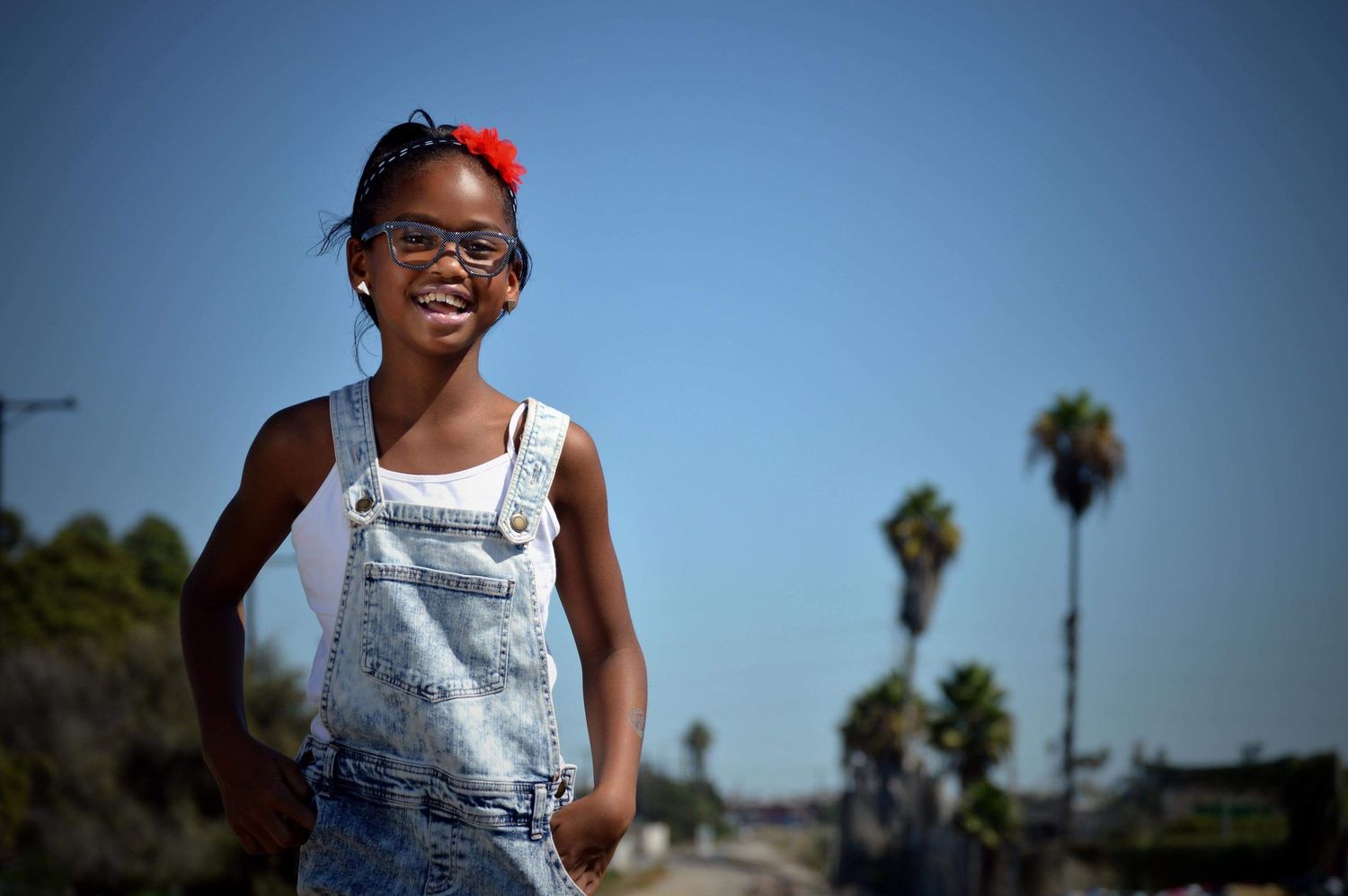Smiling young Black girl in denim overalls and glasses poses outdoors with palm trees under blue sky.