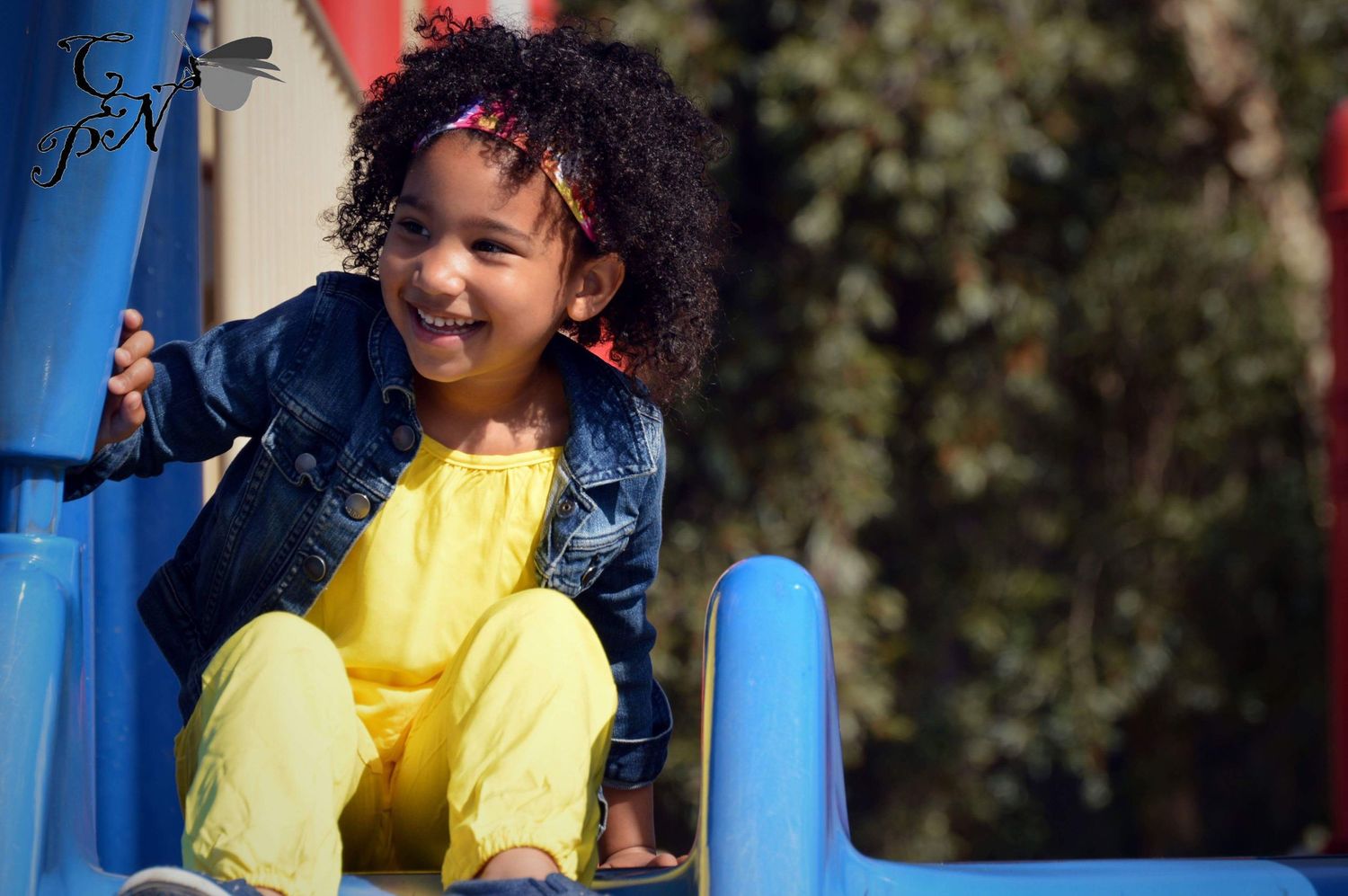 Smiling young girl with curly hair wearing yellow outfit and denim jacket playing on blue playground slide.