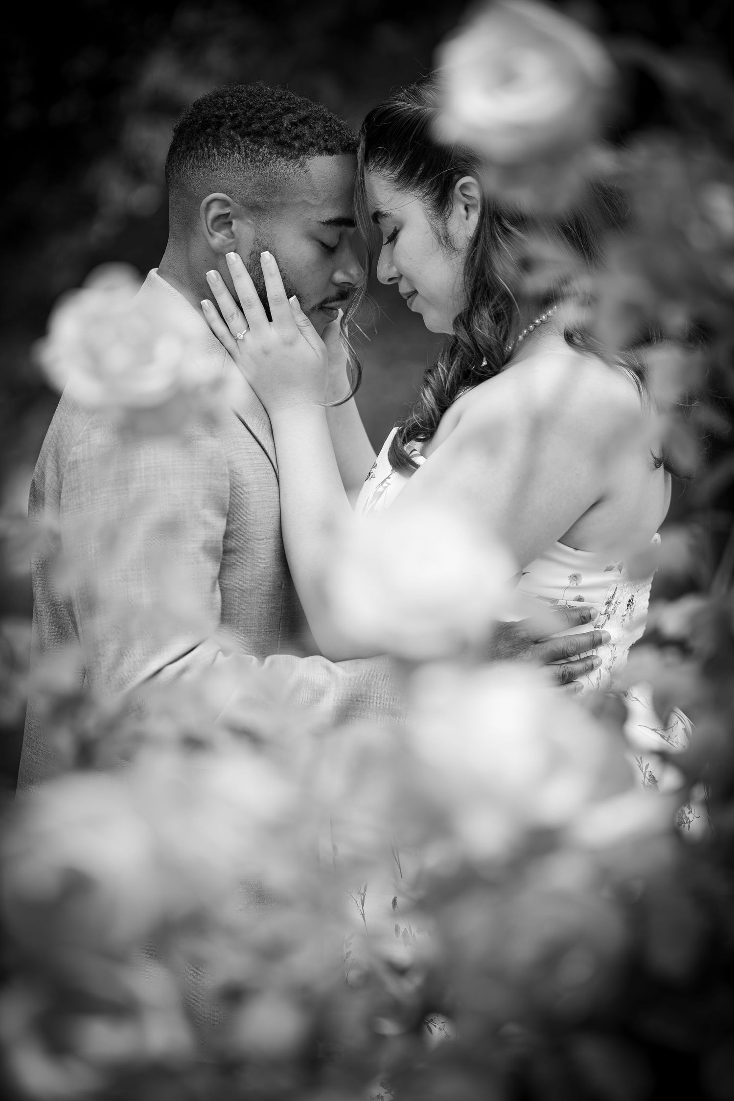 Black and white photo of a romantic couple in wedding attire sharing an intimate moment surrounded by blooming roses.