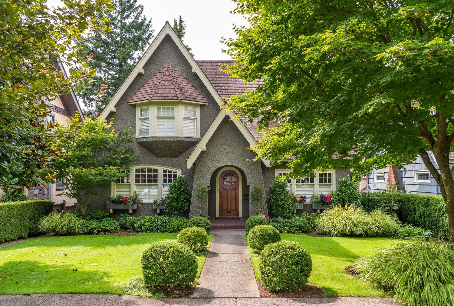 Charming Tudor-style home with lush green lawn, manicured shrubs, and tree-lined pathway leading to wooden front door.