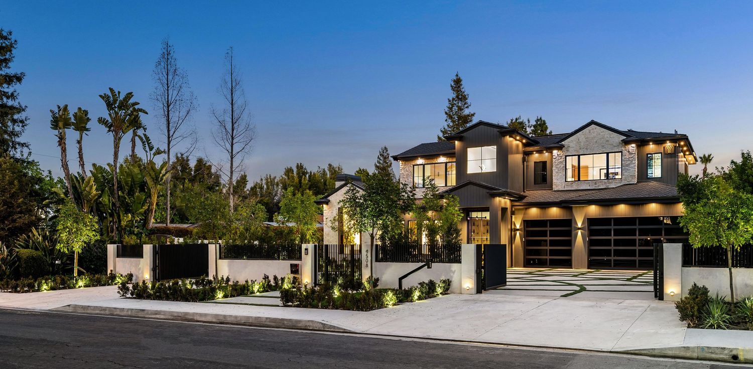 Luxury two-story modern home with dark exterior, triple garage, manicured landscaping, and palm trees at dusk in California.