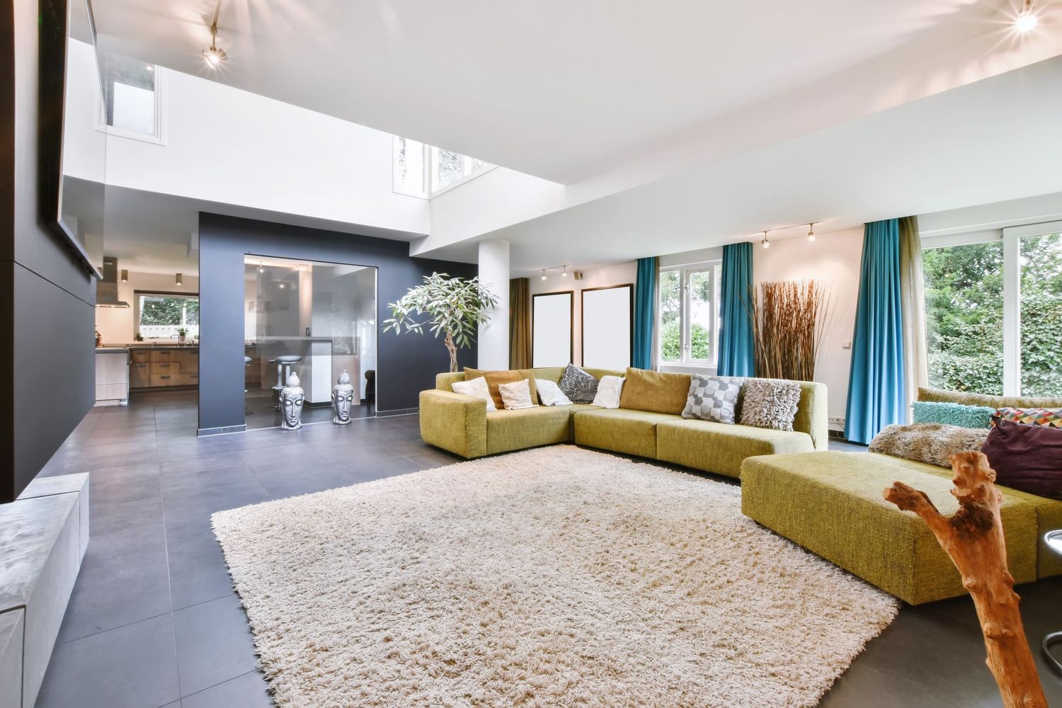 Modern open-plan living room with green sectional sofa, plush cream rug, teal curtains, and sleek kitchen in background.