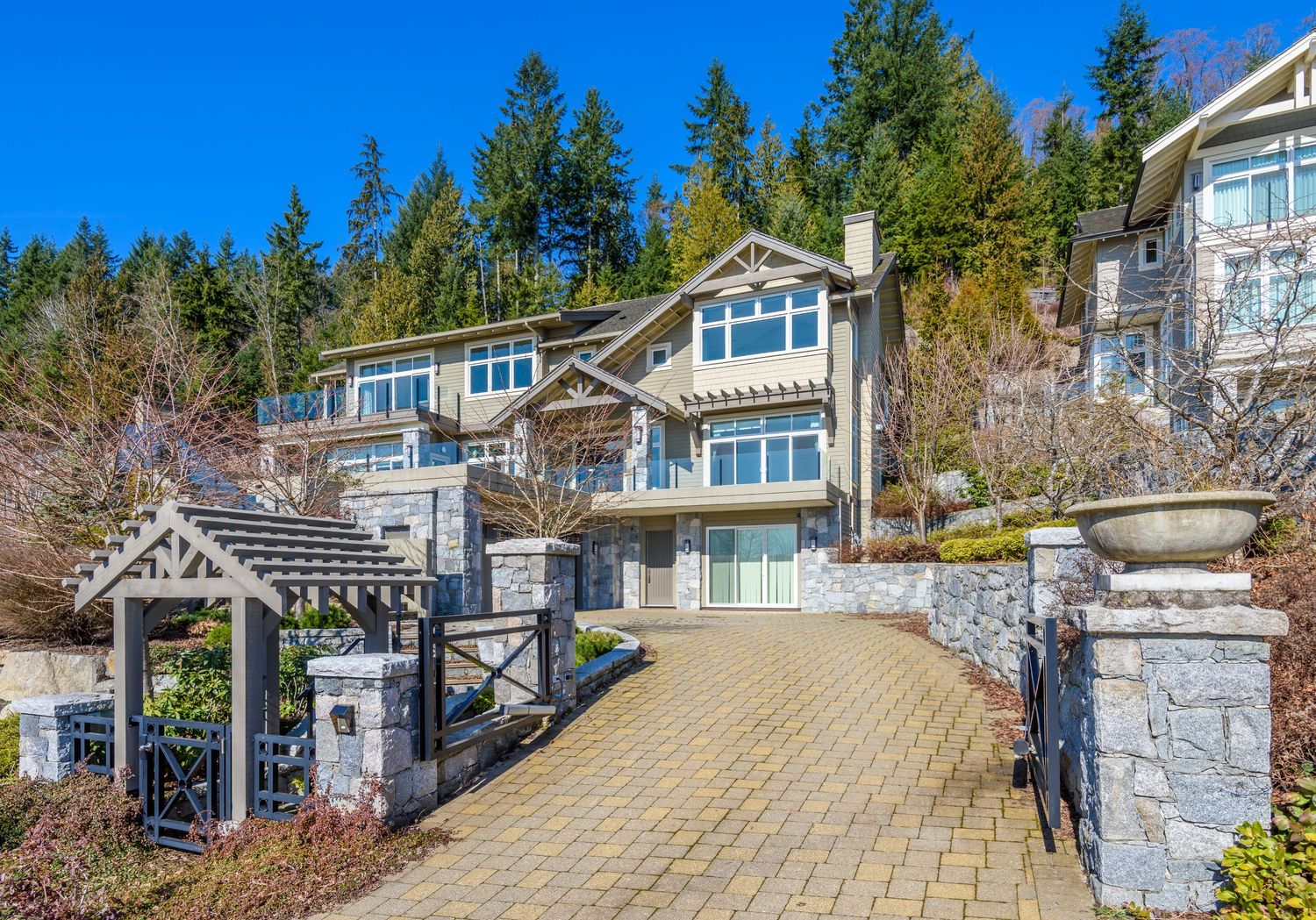 Luxury craftsman-style home with stone pillars and brick paver driveway surrounded by tall evergreen trees.