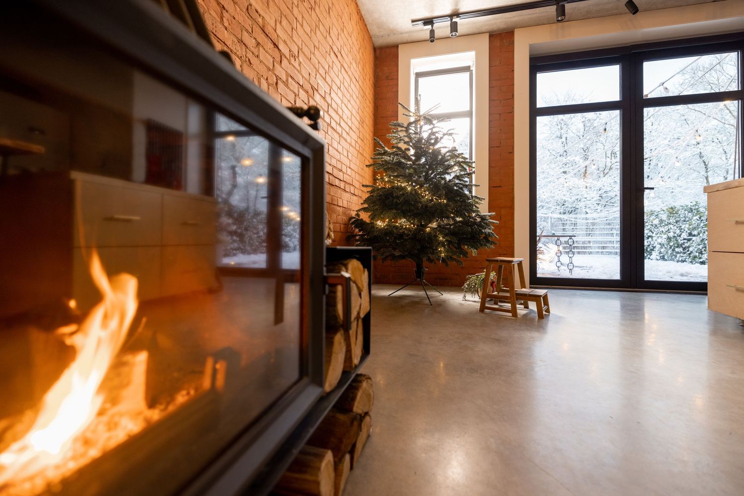 Cozy indoor fireplace with burning flames beside a decorated Christmas tree near large windows with snowy view outside.