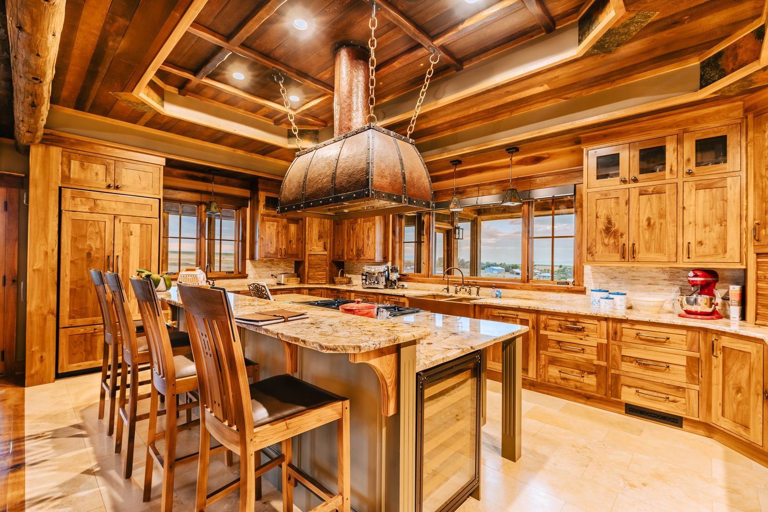 Rustic log cabin kitchen with knotty wood cabinets, granite island, bar stools, and ornate copper range hood.