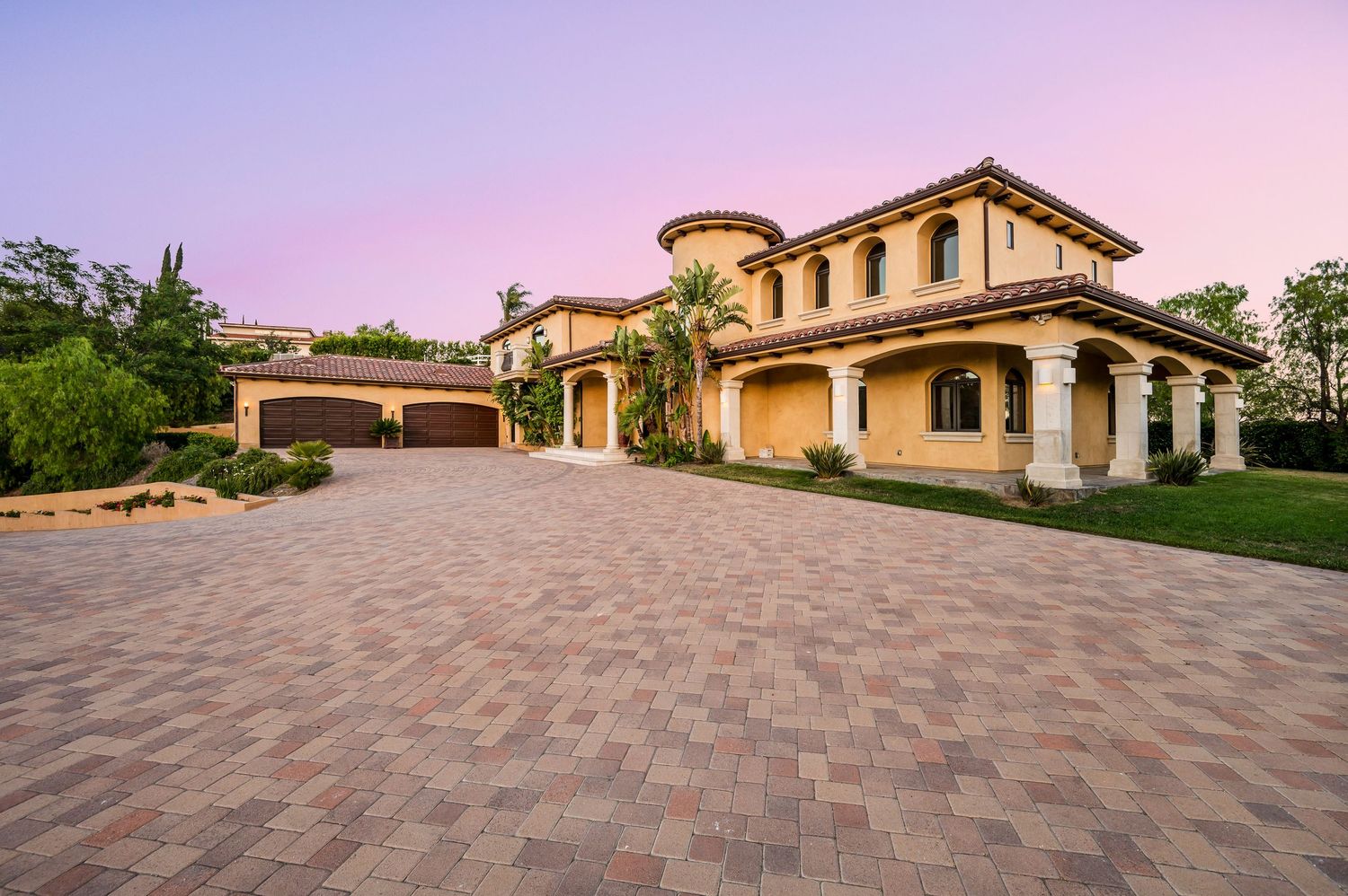 Luxury Mediterranean-style home with terracotta roof, arched columns, and elegant paver driveway at twilight.