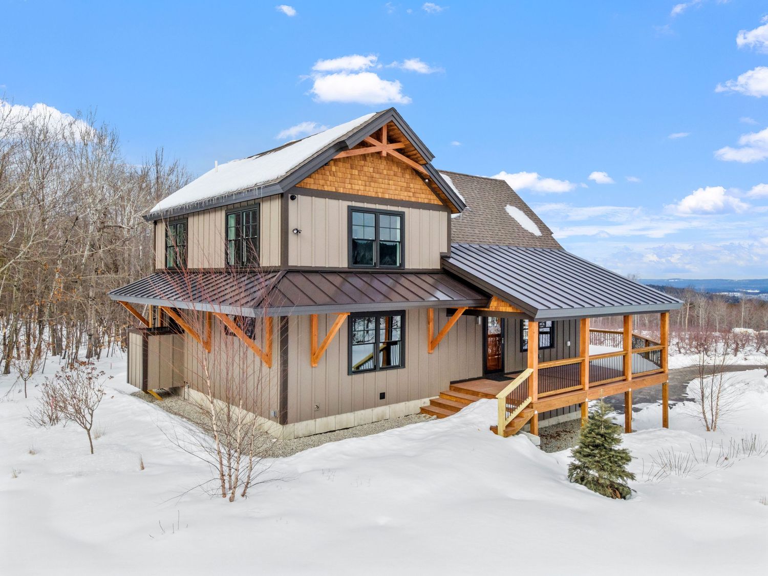 Modern mountain home with metal roof, wood accents, and wraparound deck surrounded by snow-covered landscape.