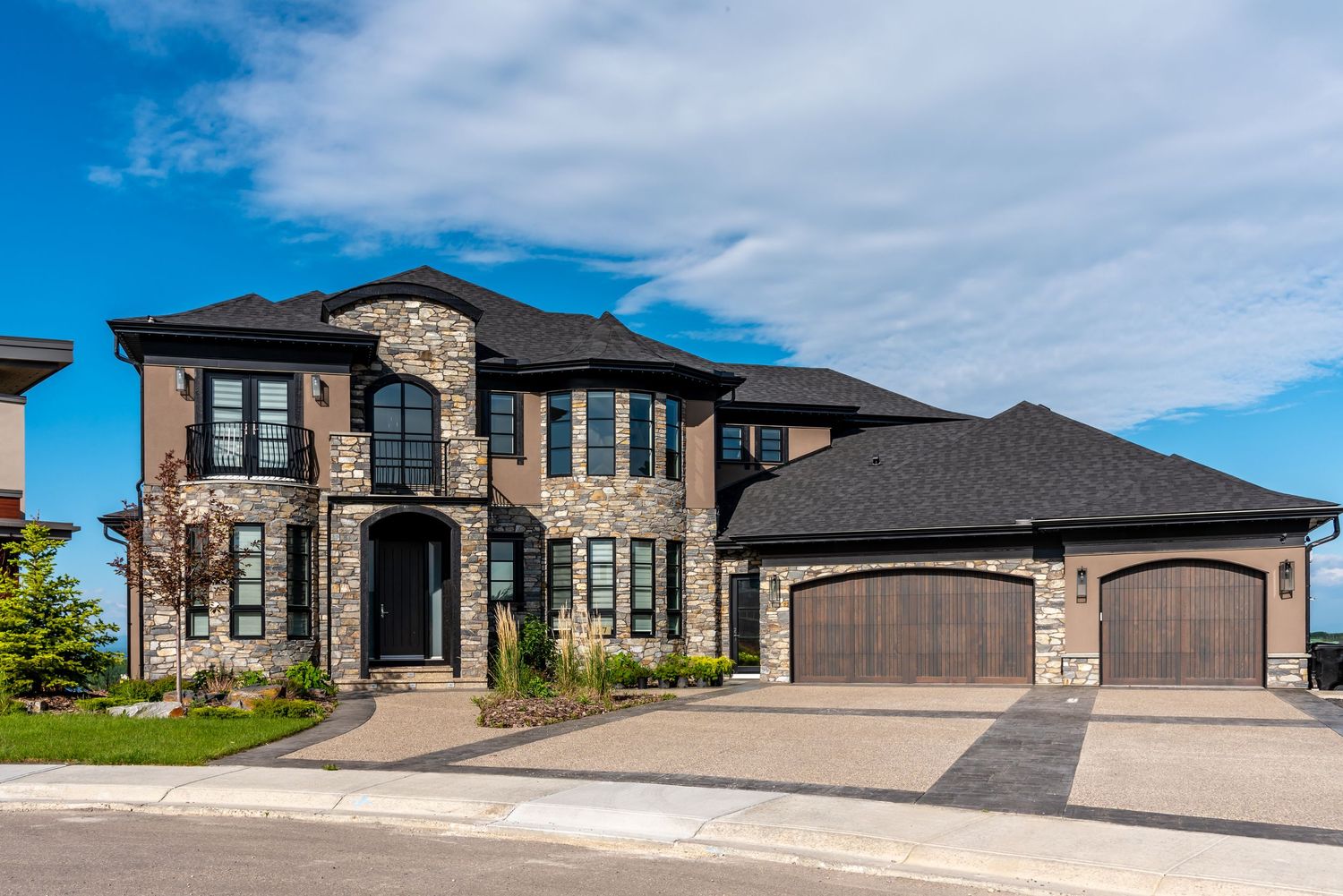 Luxury two-story stone and brick home with dark roof, three-car garage, and manicured landscaping under a blue sky.