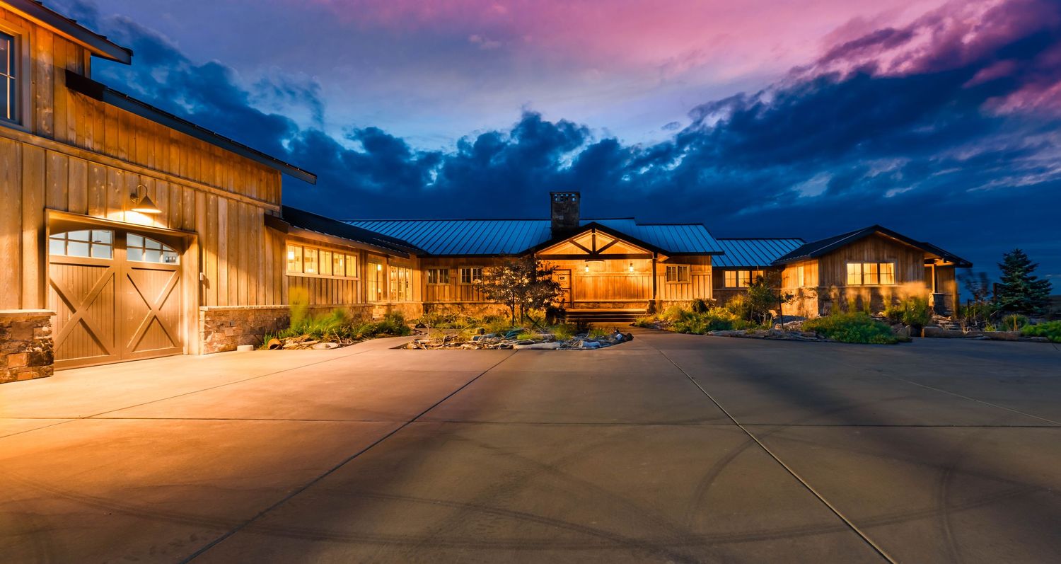 Rustic ranch-style home with warm lighting at dusk, featuring wooden barn doors and a blue metal roof under dramatic clouds.