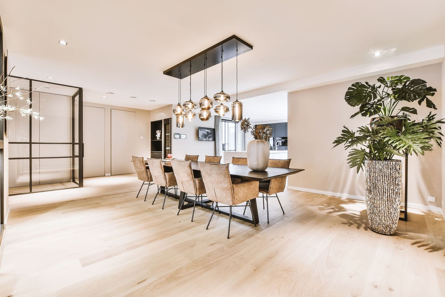 Modern dining room with long wooden table, pendant lights, light hardwood floors, and a large tropical plant in a vase.