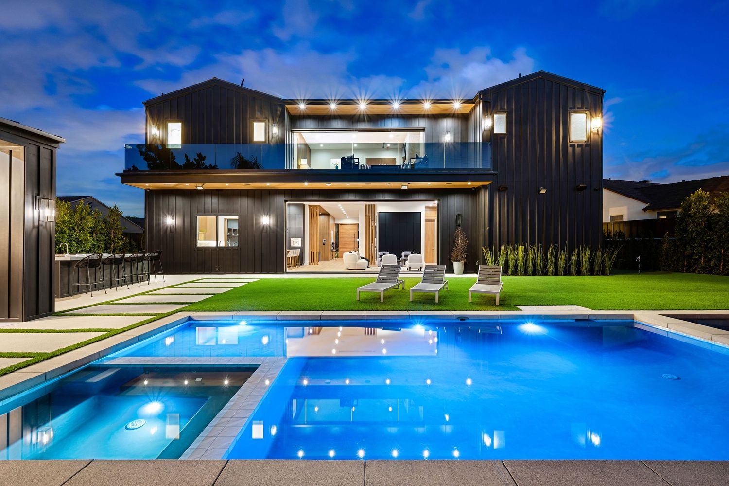 Modern black two-story home with illuminated pool, lush lawn, and outdoor seating at dusk.
