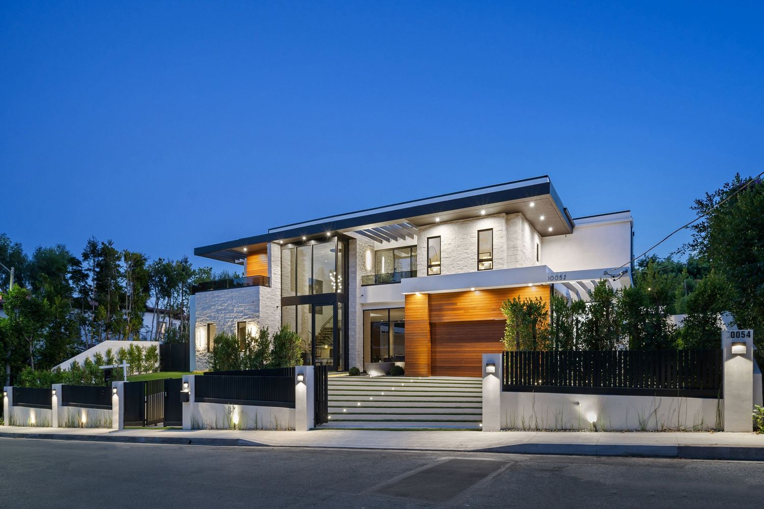 Modern two-story luxury home with white exterior, wood garage accents, and elegant landscape lighting at dusk.