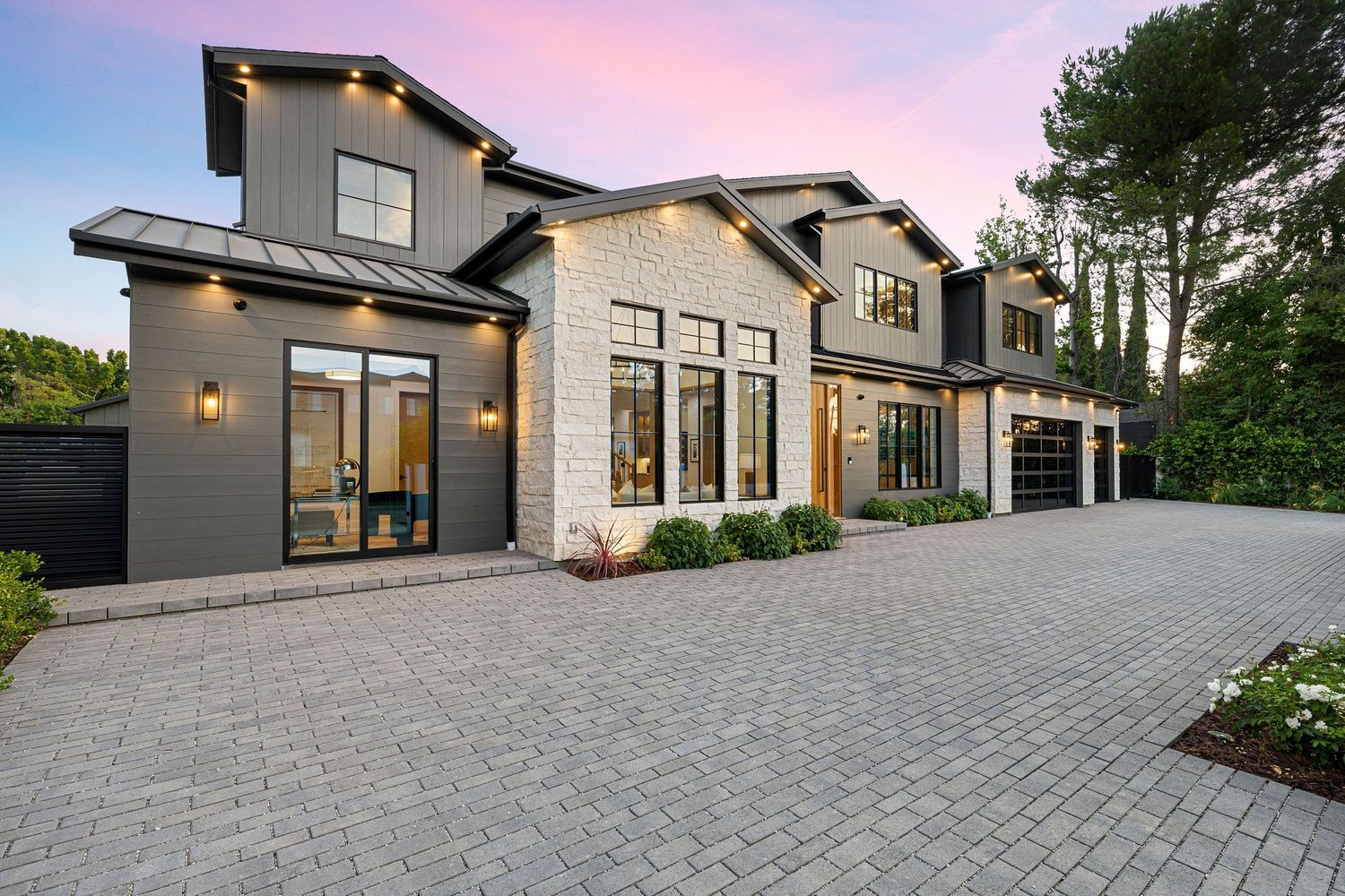 Modern farmhouse exterior with dark siding, stone accents, 3-car garage, and paver driveway at dusk.
